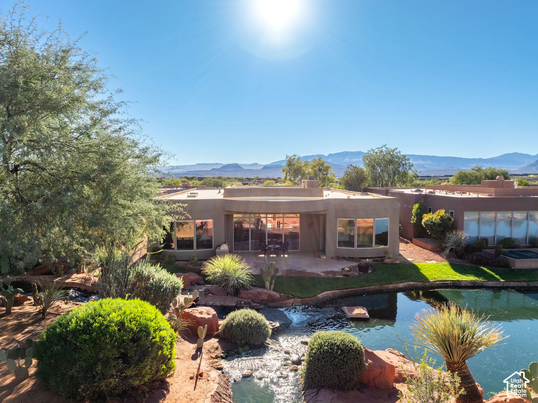 Stucco-exterior residence featuring expansive windows, a rock-lined water feature, terraced landscaping, and a mountain backdrop