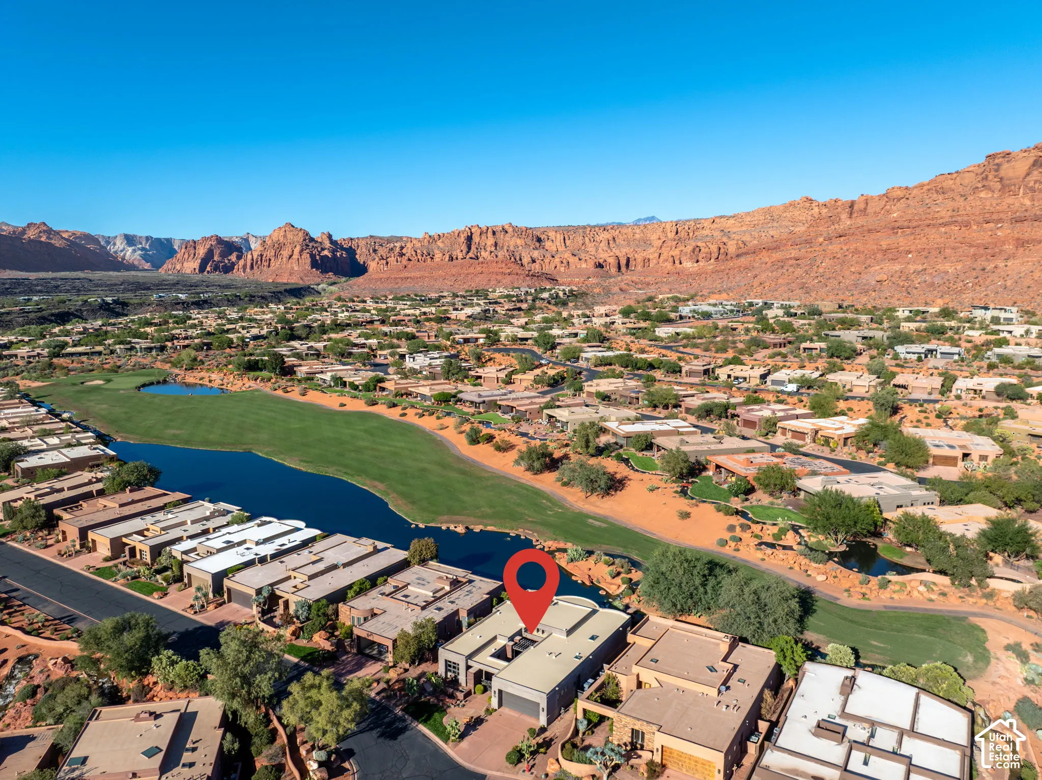 Aerial perspective of a multi-level flat-roof residence featuring exterior courtyards and a paved driveway, situated in a desert landscape with sandstone mountain topography and a golf course water feature