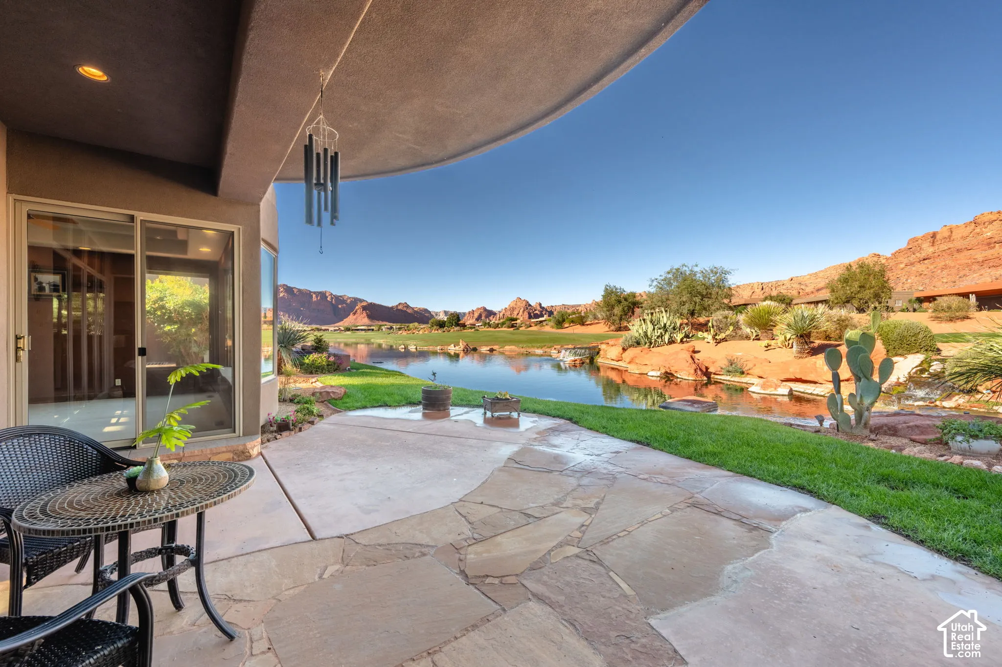 Covered stone patio featuring flagstone paving, integrated recessed lighting, sliding glass door, and immediate proximity to a pond and red rock mountain backdrop