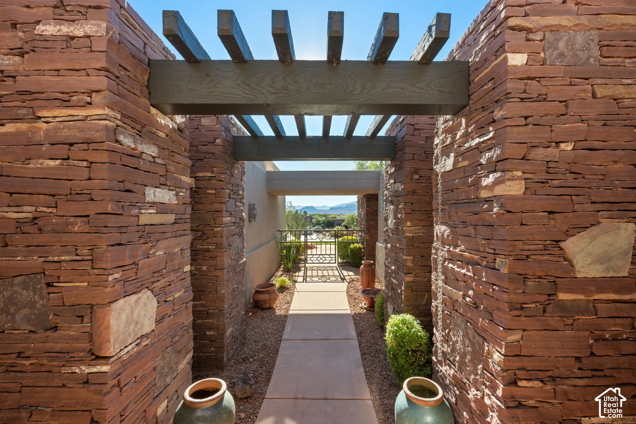 Entryway featuring stacked stone walls, a dark-finished wood pergola, concrete paving, and a decorative metal gate