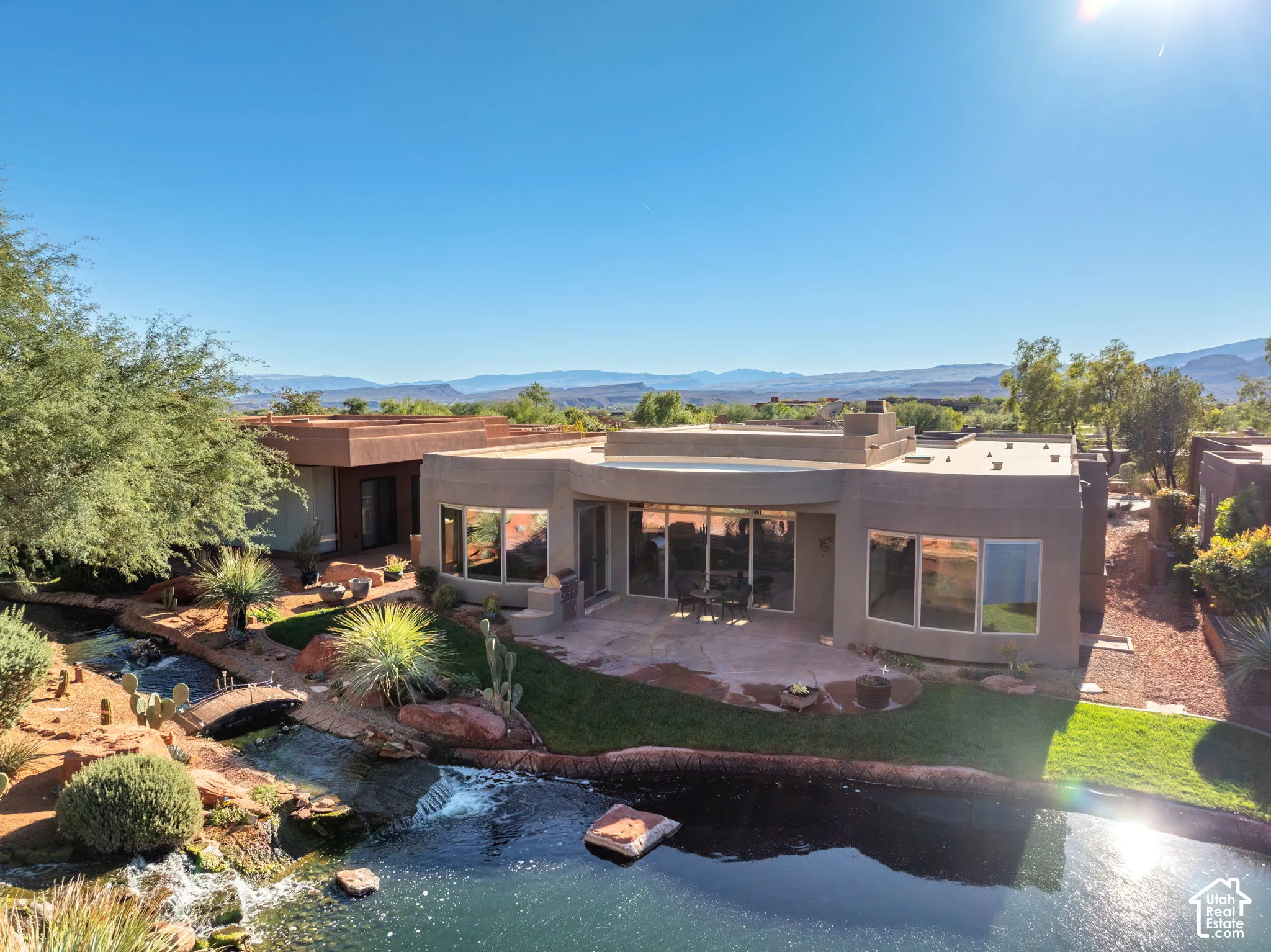 Stucco exterior featuring floor-to-ceiling windows, stone patio, custom water feature with stone-lined pond, and xeriscaped grounds with mature desert vegetation