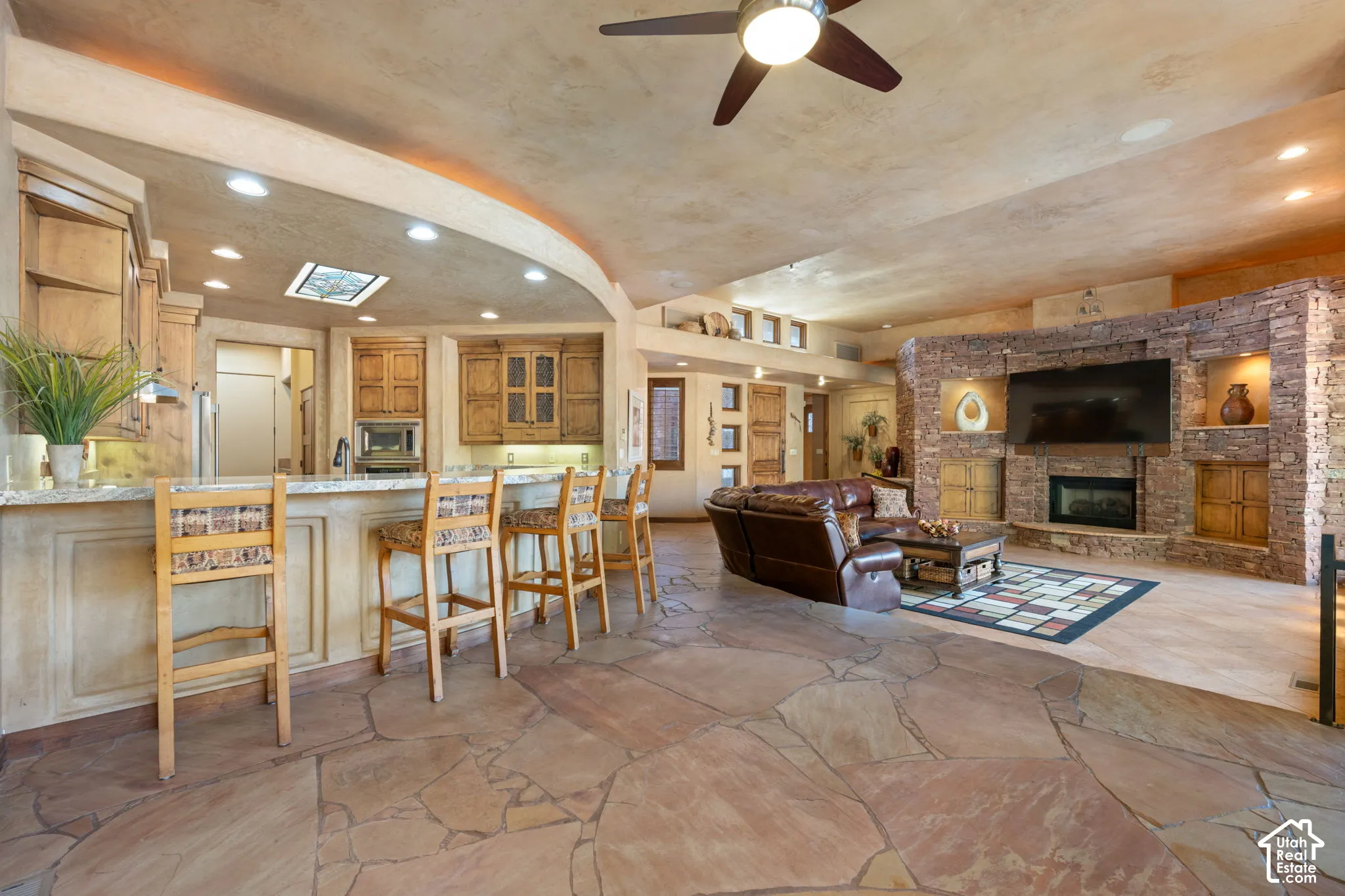 Open-concept interior featuring natural stone flooring, arched kitchen island with stone-surface countertop, floor-to-ceiling stone fireplace surround, and recessed lighting