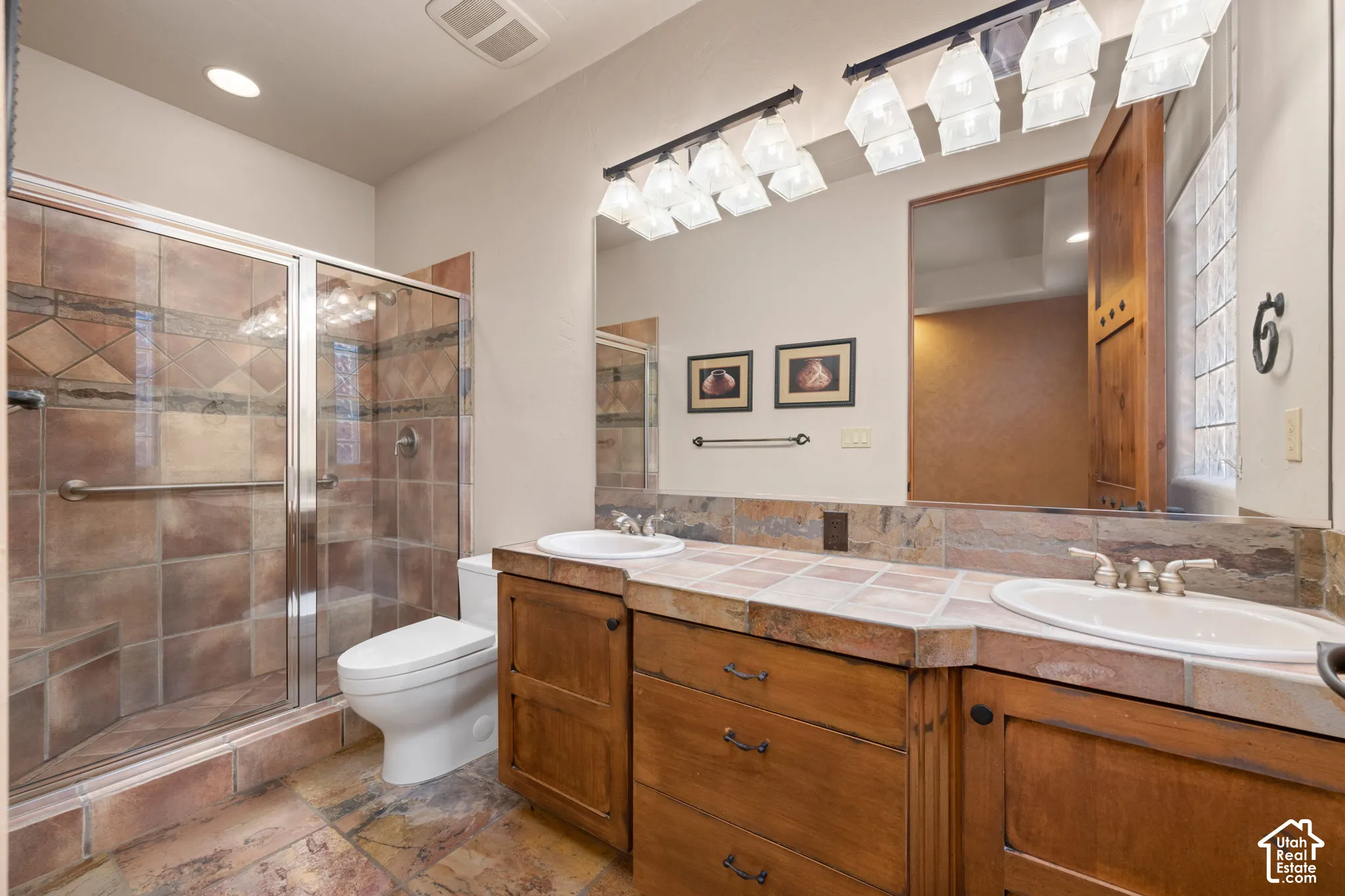 Bathroom featuring a dual-sink vanity with wood cabinetry and tile-surfaced countertop, stone-tile flooring, and a walk-in shower with stone-tile surround and glass door