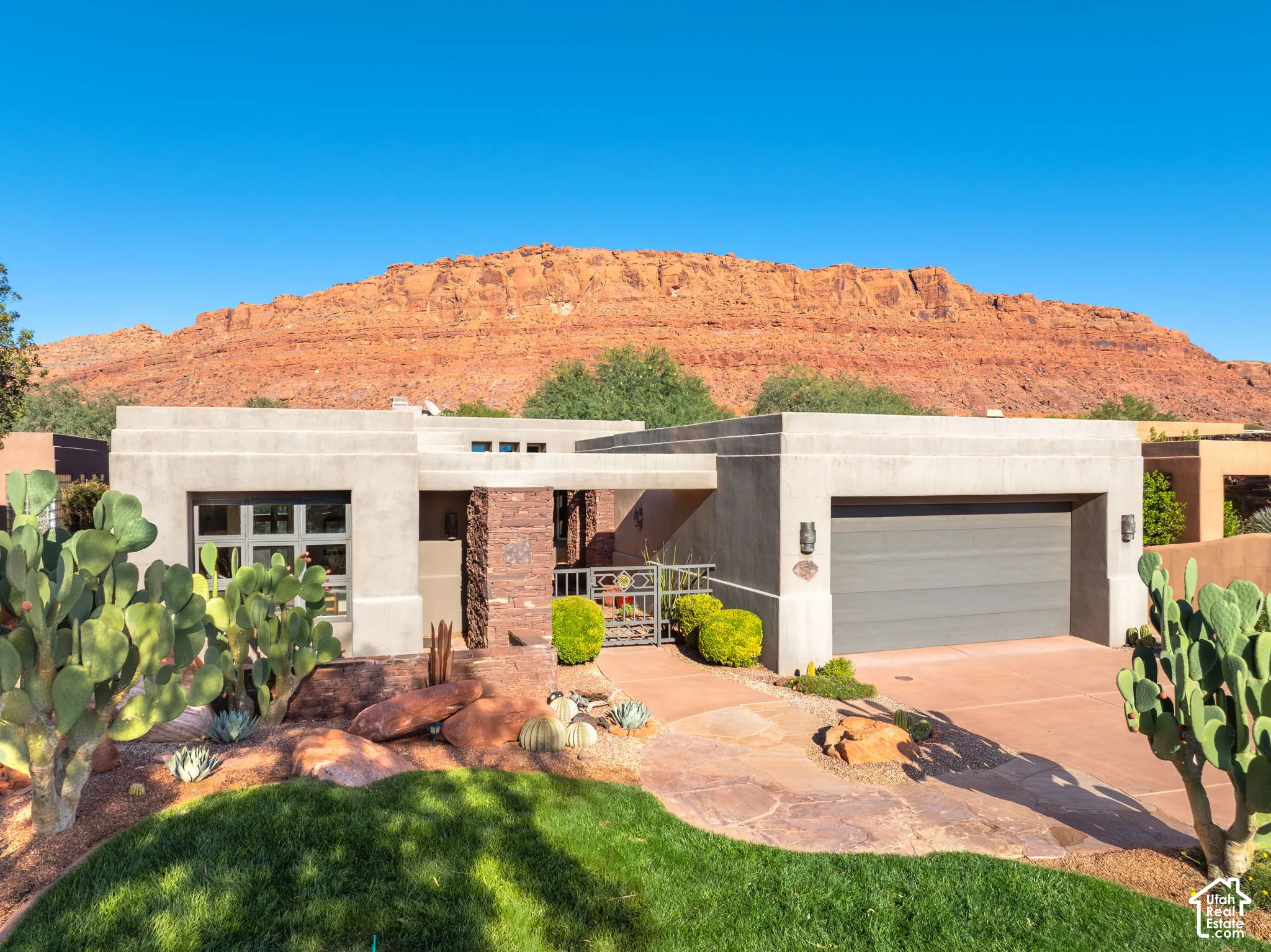 Stucco exterior featuring stacked stone accents, flat roofline, attached multi-car garage, and desert landscaping with mature cactus specimens against a red rock backdrop
