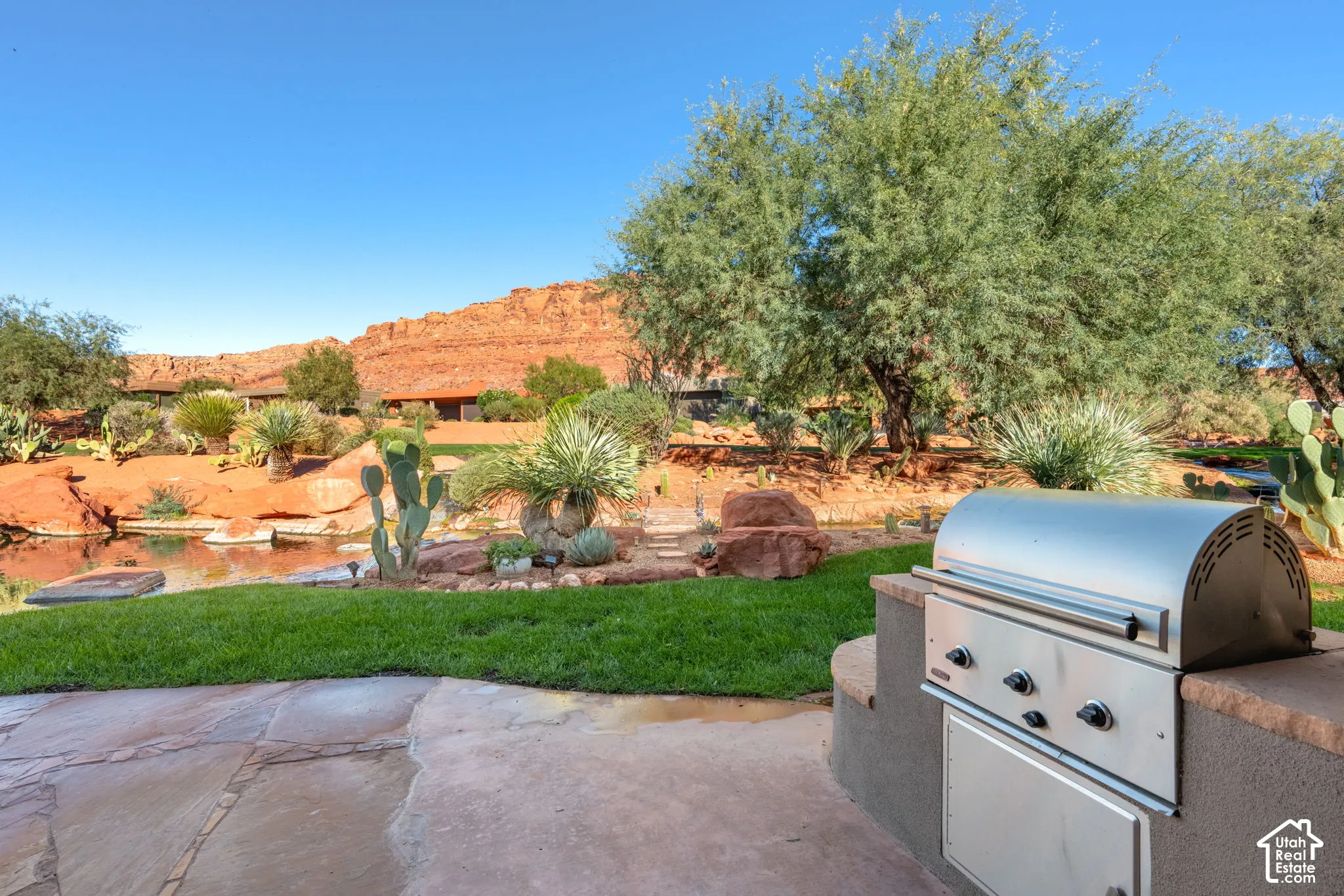 Outdoor patio featuring a built-in stainless steel grill, natural stone paving, manicured lawn, water feature, and desert landscaping set against a rock formation backdrop