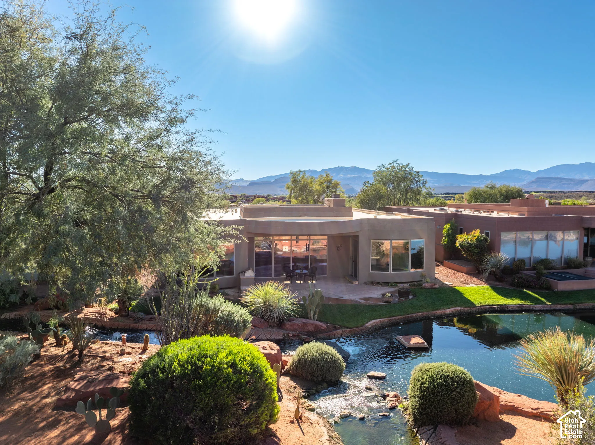 Stucco exterior featuring floor-to-ceiling glass windows, a landscaped water feature with natural stone edging, mature desert vegetation, and a mountain backdrop