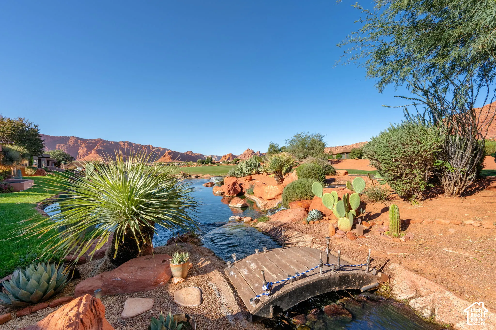 Landscaped grounds featuring a stone-lined water feature, wooden footbridge, native desert vegetation, and red rock mountain backdrop
