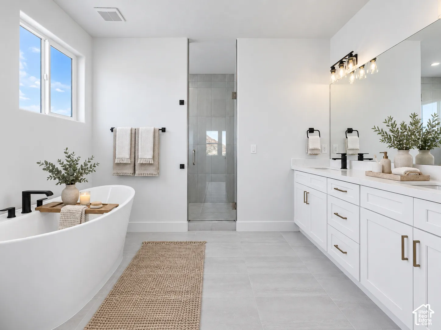 Freestanding soaking tub with matte black fixtures, white shaker-style dual vanity with solid surface countertops, expansive wall-to-wall mirror, and glass-enclosed shower with tile surround