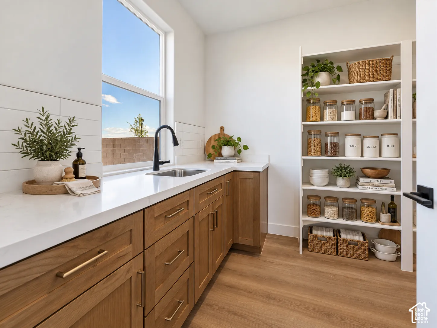Natural wood cabinetry featuring metal hardware, white stone countertops, undermount sink with a matte black faucet, horizontal white tile backsplash, and built-in floor-to-ceiling white shelving