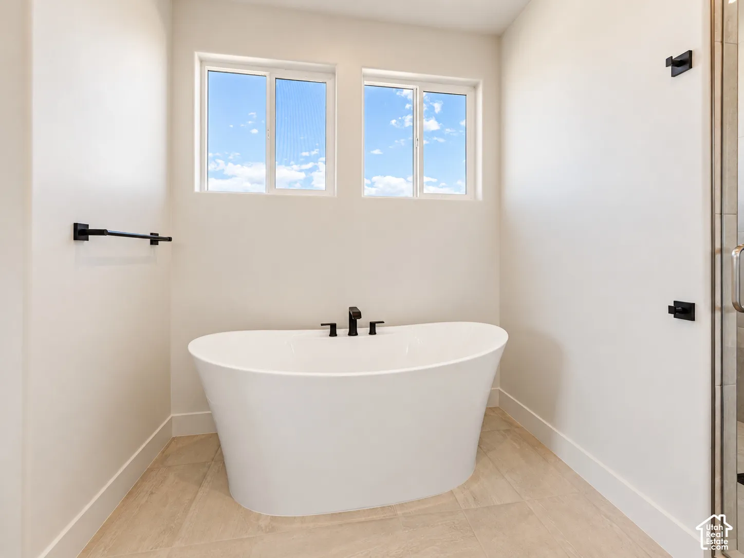 Freestanding white soaking tub with matte black deck-mounted faucet, dual windows, and light-toned tile flooring
