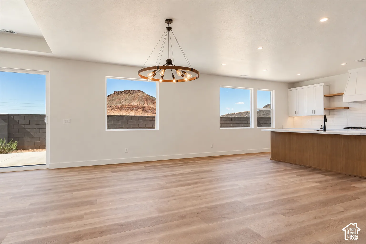 Open concept interior featuring light wood-finish flooring, a wrought-iron wagon wheel chandelier, recessed lighting, white cabinetry with a wood-accented kitchen island, and multiple windows showcasing a mountain backdrop