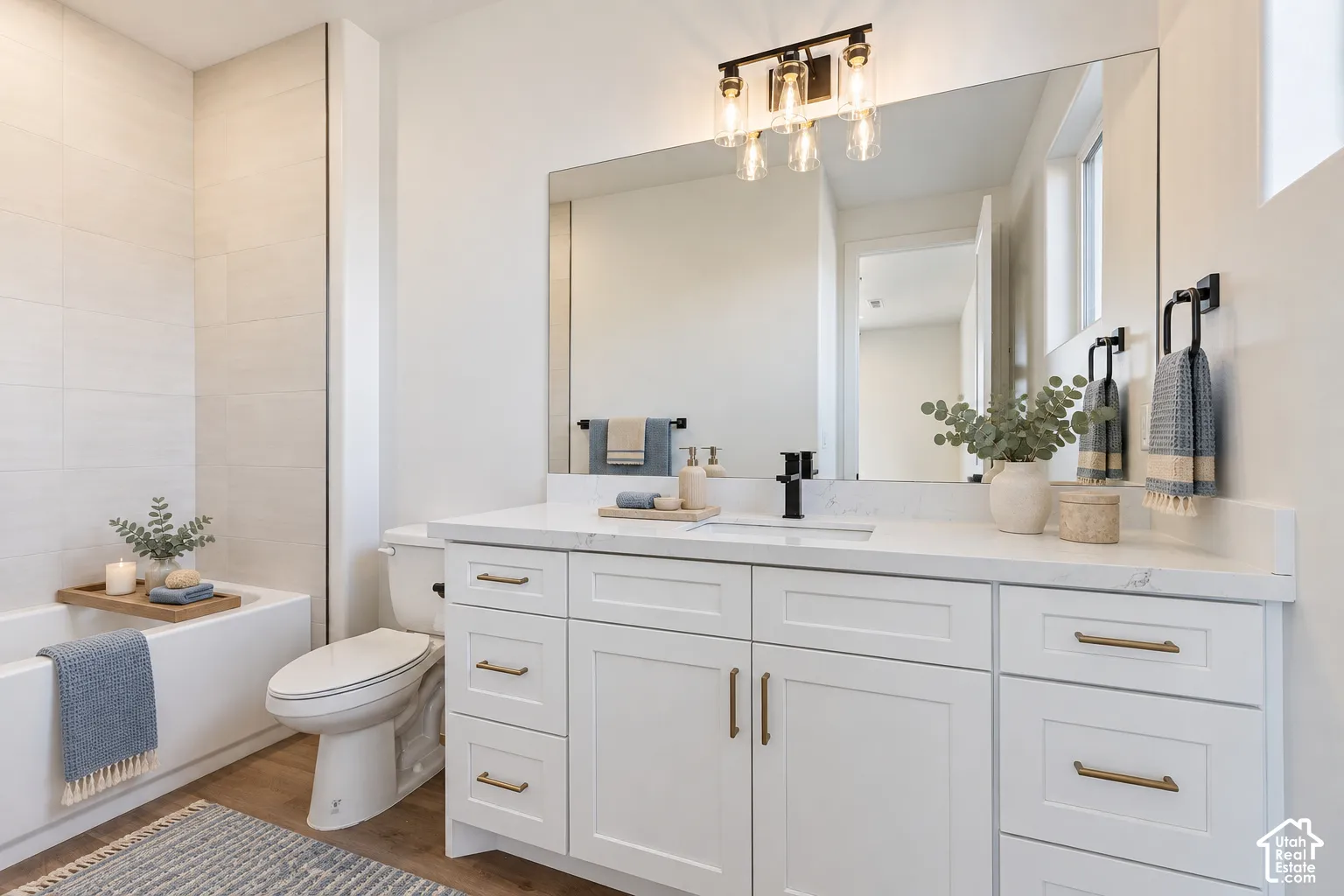 Bathroom featuring a white shaker-style vanity with stone-finish countertop, matte black faucet, wall-to-wall mirror, and a bathtub with tiled surround