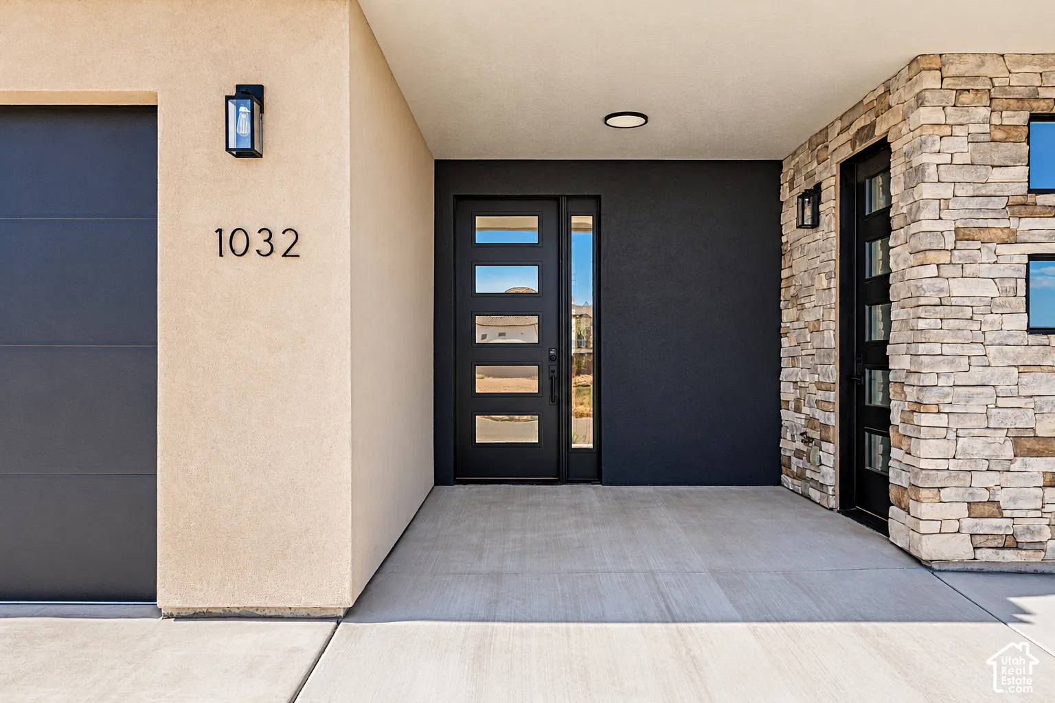 Modern entryway featuring a multi-panel door with glass inserts, stacked stone veneer siding, smooth stucco exterior finish, recessed ceiling lighting, and matte black wall-mounted fixtures