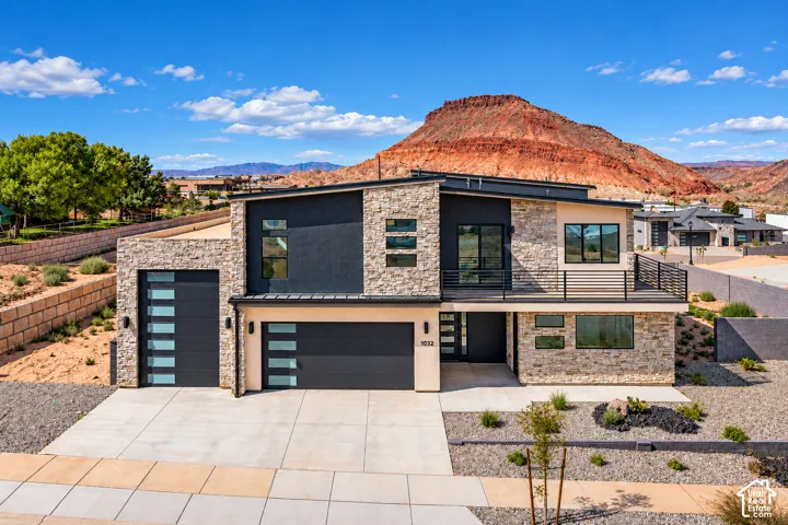 Contemporary multi-level exterior featuring a stone veneer facade, dark stucco accents, a shed-style roofline, a second-story balcony with metal railings, and integrated garage doors with horizontal glass panels