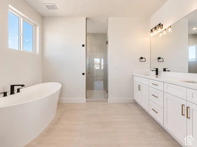 Freestanding soaking tub with matte black fixtures, white shaker-style cabinetry with gold-tone hardware, quartz countertop with dual integrated sinks, glass-enclosed shower with tiled surround, and large-format tile flooring