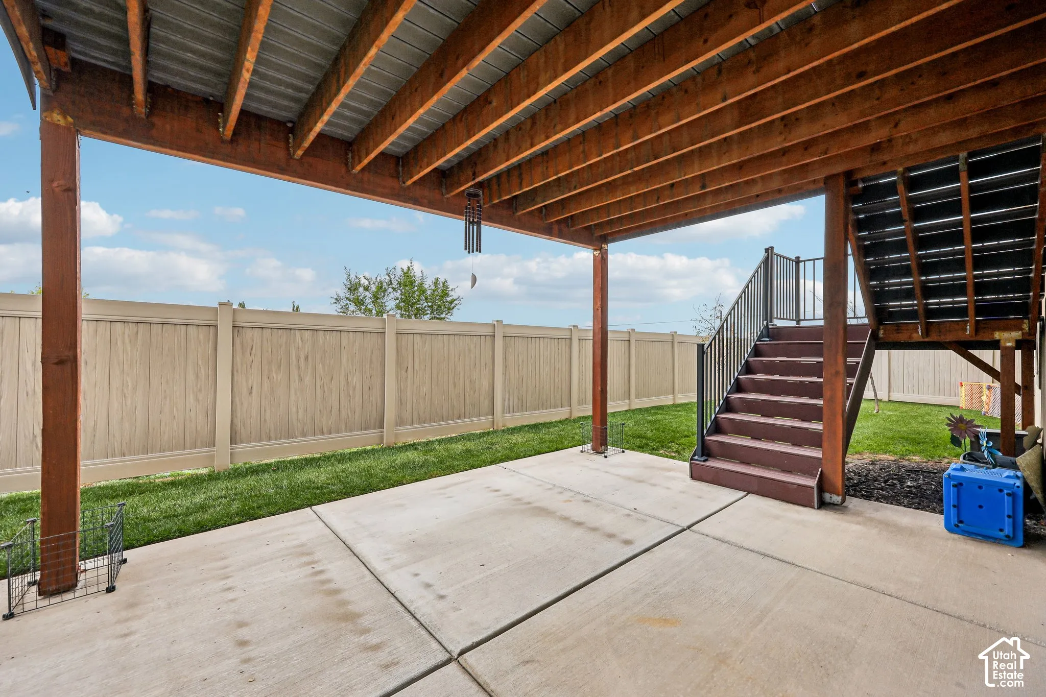 Covered concrete patio featuring exposed timber ceiling joists, wood-finish stairs with metal railings, and a fenced backyard