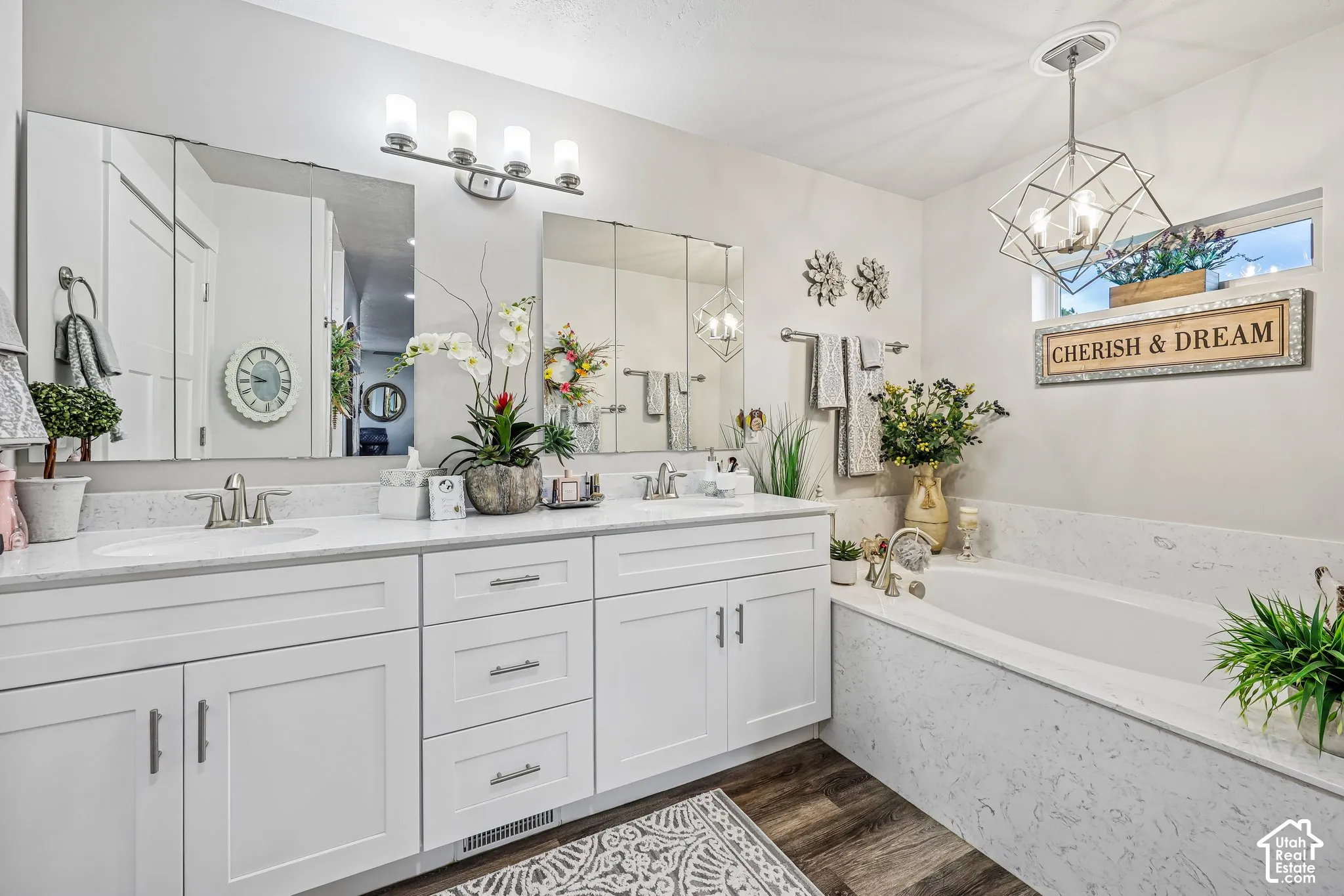 Double vanity with white cabinetry, stone-surfaced countertops, recessed medicine cabinets, and geometric pendant lighting, complemented by a built-in soaking tub with matching stone surround and wood-finish flooring