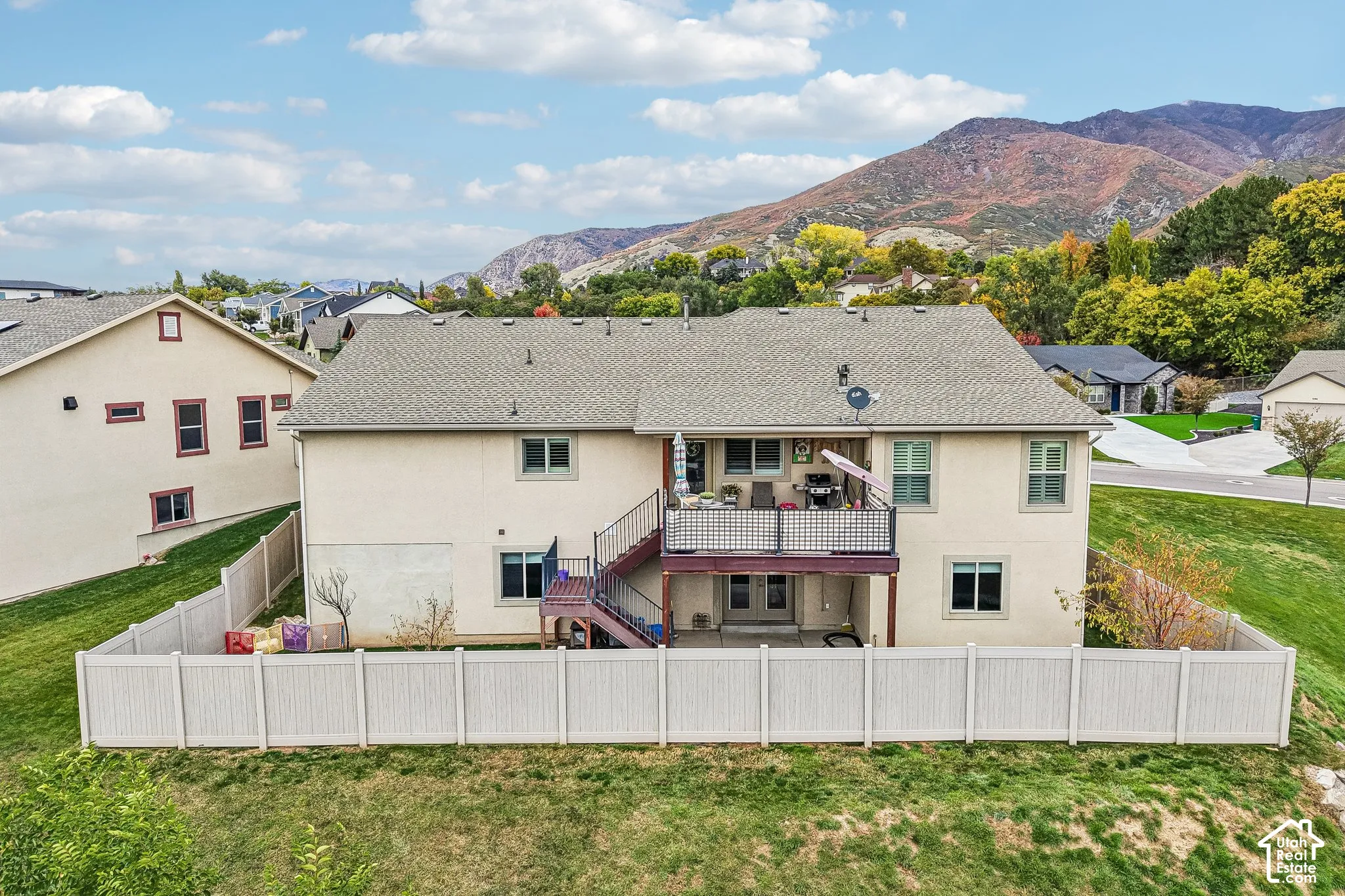 Two-story stucco exterior featuring a raised deck with metal railing, an exterior staircase, a concrete patio, and a perimeter privacy fence, all set against a mountain backdrop