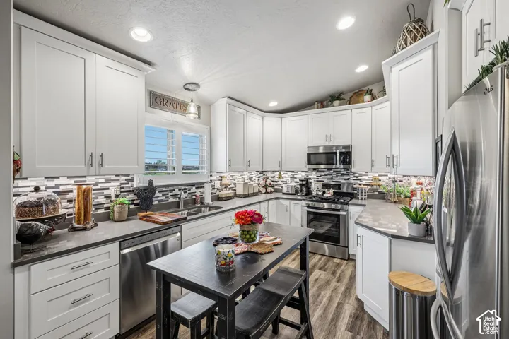 Kitchen featuring white shaker-style cabinetry, solid-surface countertops, a multicolored mosaic tile backsplash, stainless steel appliances, and wood-finish flooring