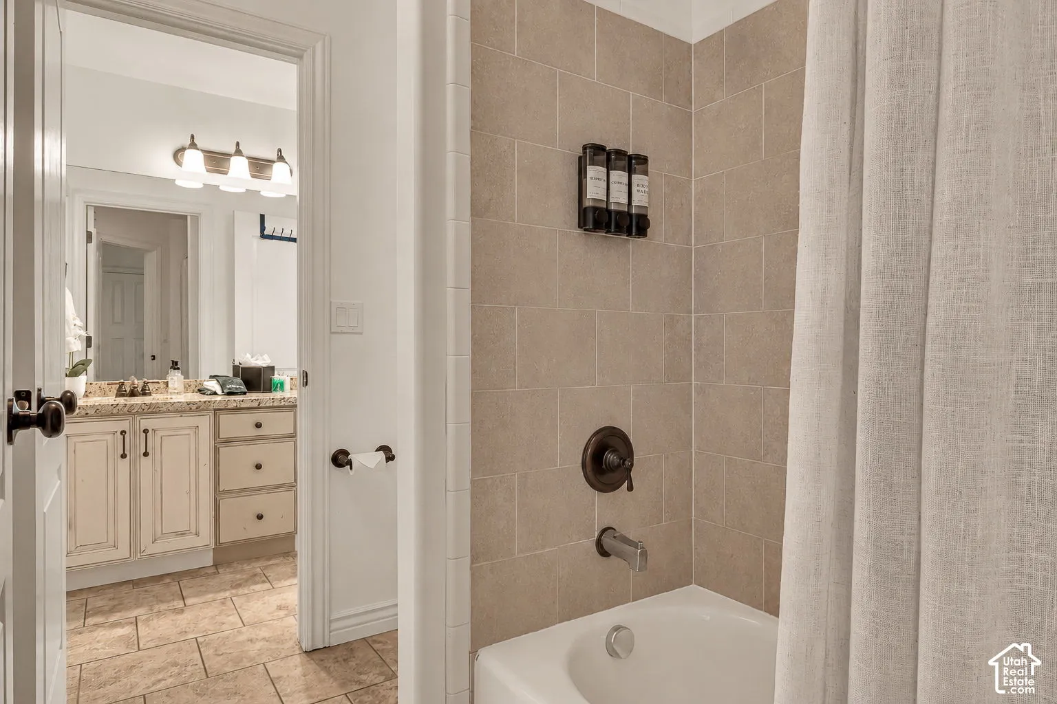 Bathtub featuring neutral-toned tile surround with bronze-finish hardware, paired with a vanity cabinet containing stone-surface counters, multi-drawer storage, and a four-bulb light fixture