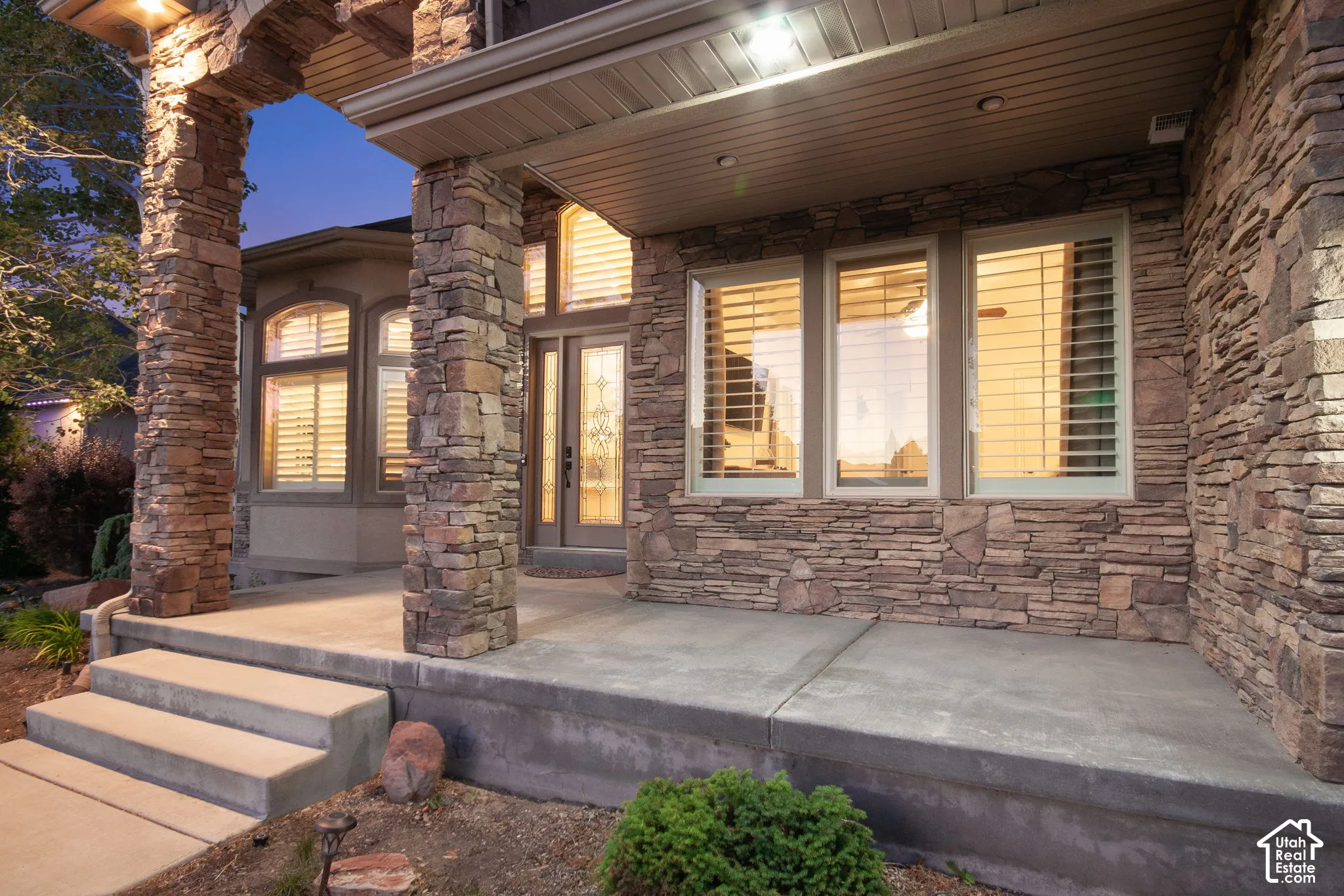 Covered porch featuring stacked stone siding, structural stone columns, concrete steps, and glass-paneled entry door with sidelights
