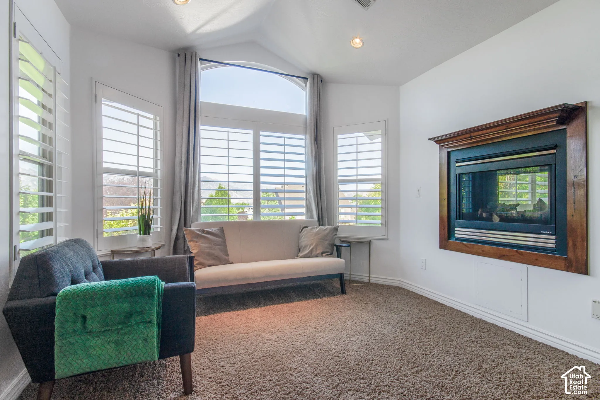 Vaulted ceilings featuring recessed lighting, large arched window, wall-mounted fireplace with wood-finish surround, and plantation shutters