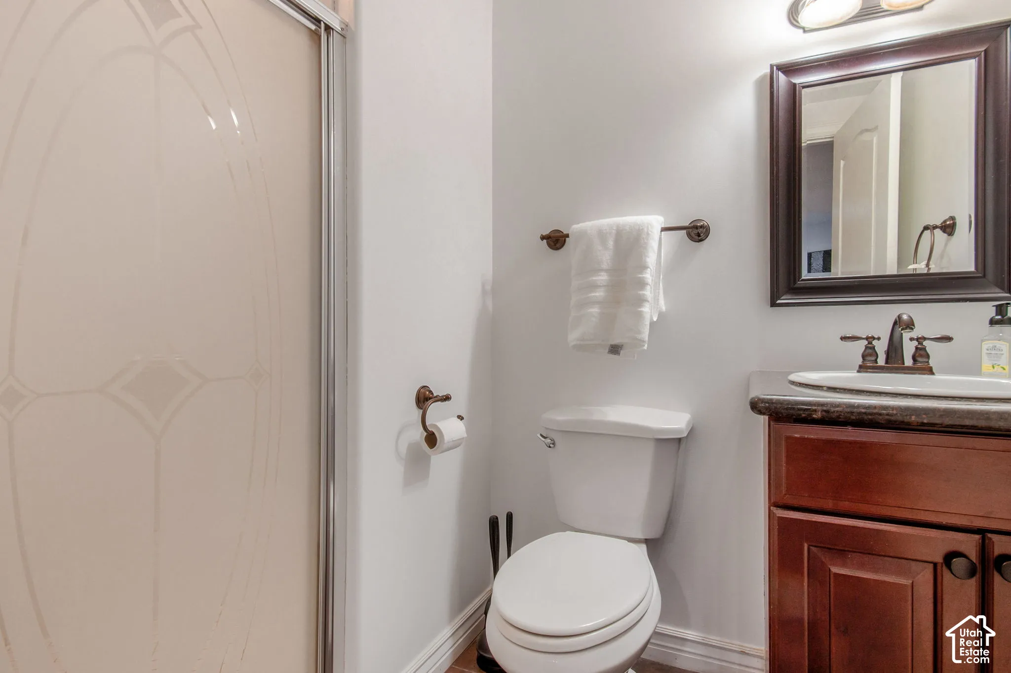Bathroom featuring a wood-finish vanity with a stone-surface countertop, an oval under-mount sink with bronze-tone hardware, a framed mirror, and a walk-in shower enclosure with glass doors