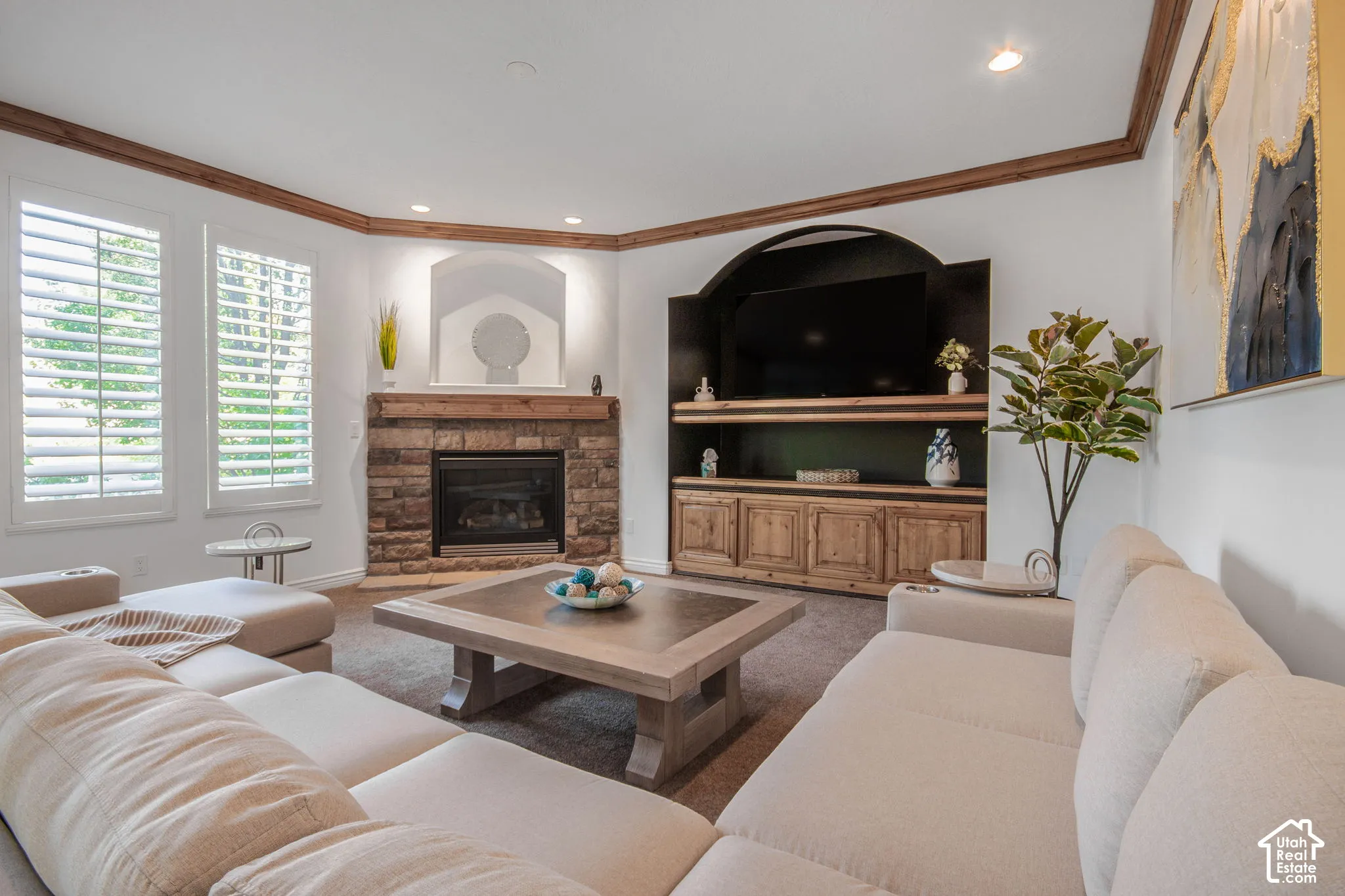 Corner stone-surround fireplace with wood mantel, built-in wooden cabinetry, recessed arched niche, crown molding, and plantation shutters