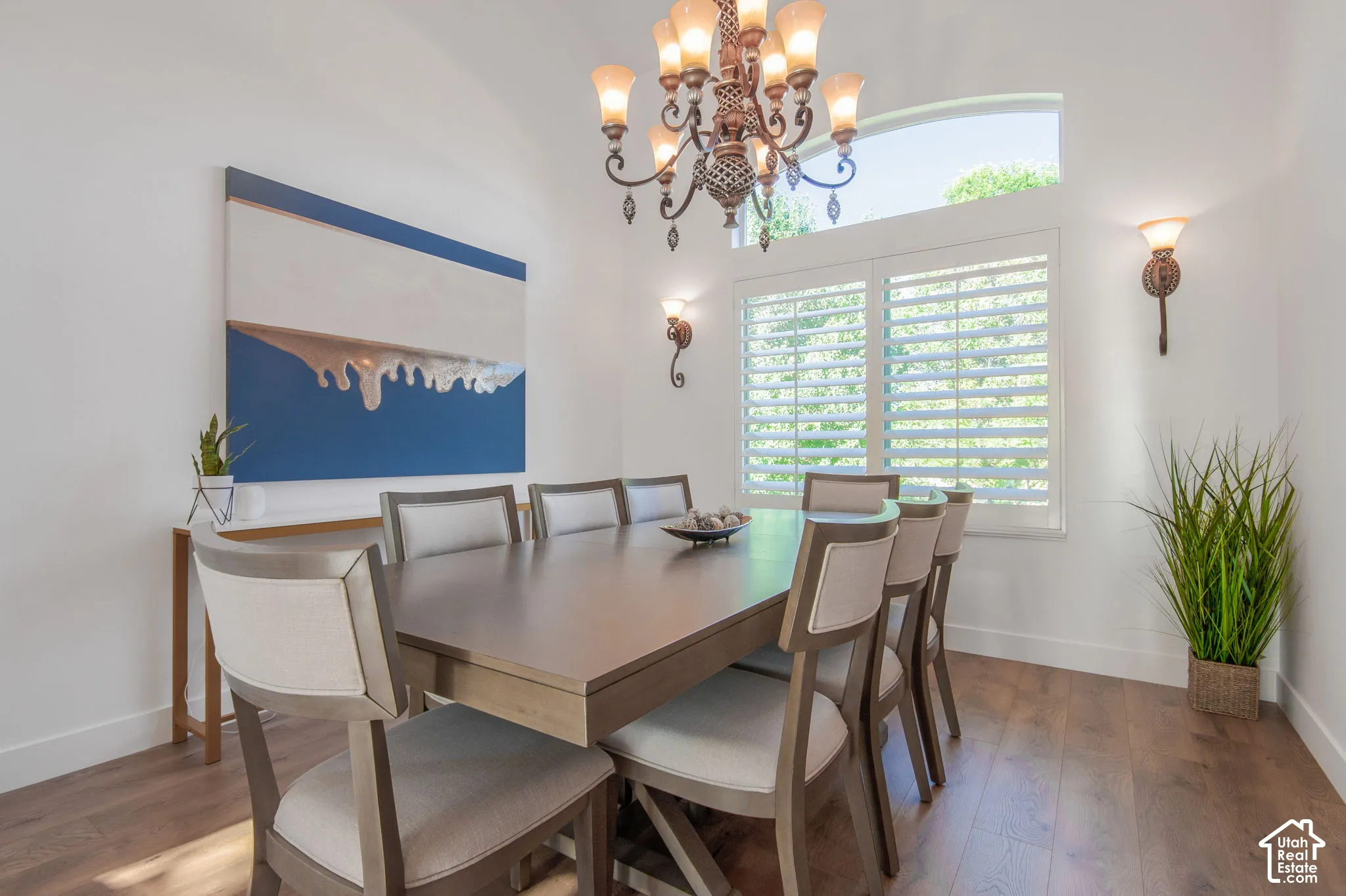 Vaulted ceiling featuring a tiered chandelier, custom plantation shutters, arched transom window, wood-finish flooring, and decorative wall-mounted light fixtures