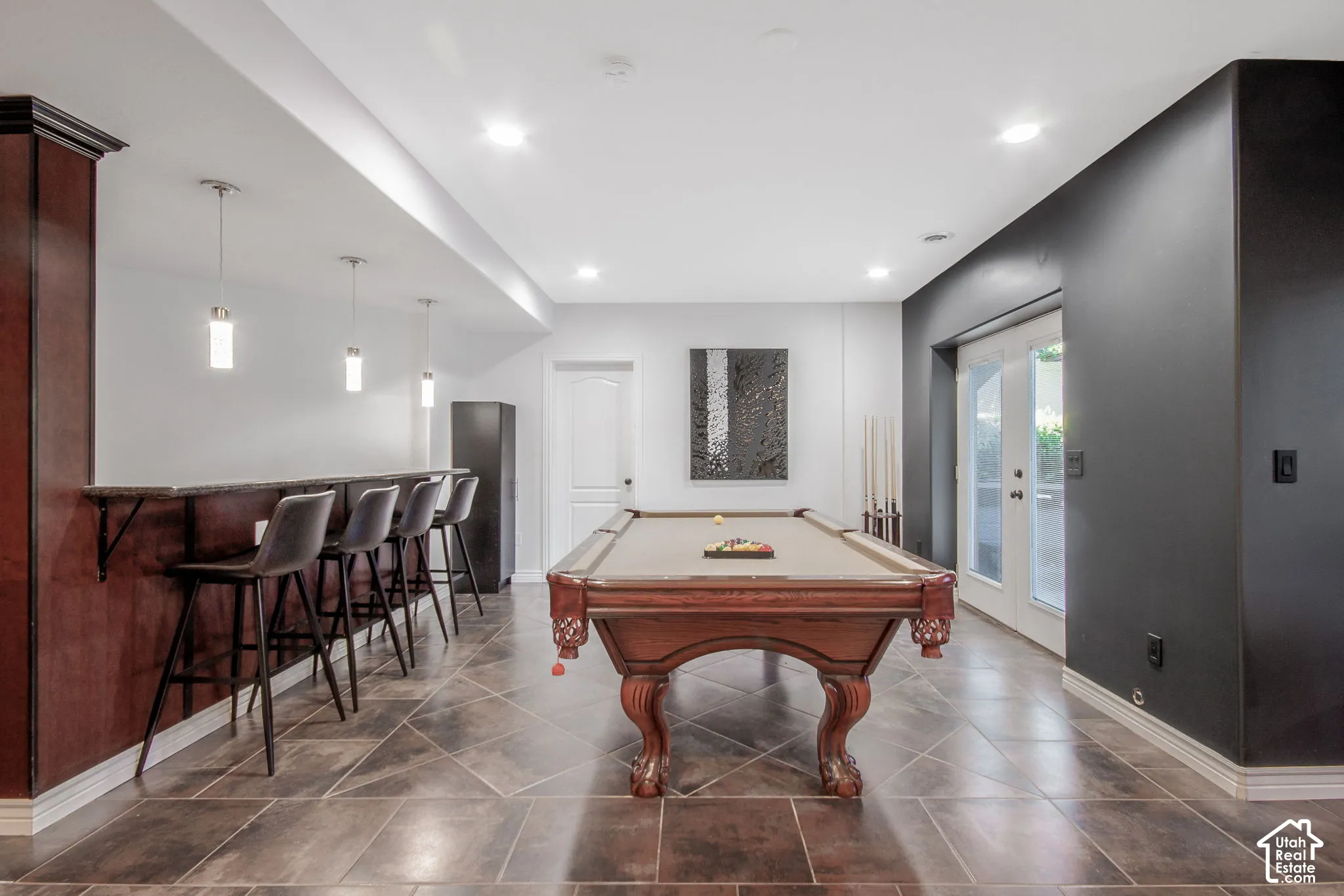Built-in wet bar featuring dark wood cabinetry, integrated bar counter, recessed lighting, and pendant fixtures set against large-format tile flooring and dual glass-panel exterior doors