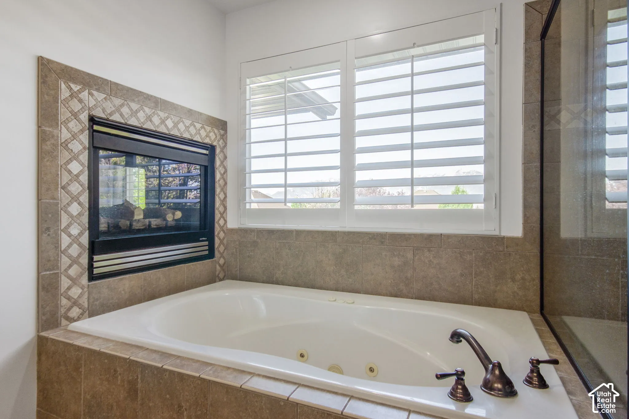 Jetted soaking tub with bronze-finish fixtures, inset fireplace with decorative tile surround, and plantation shutters