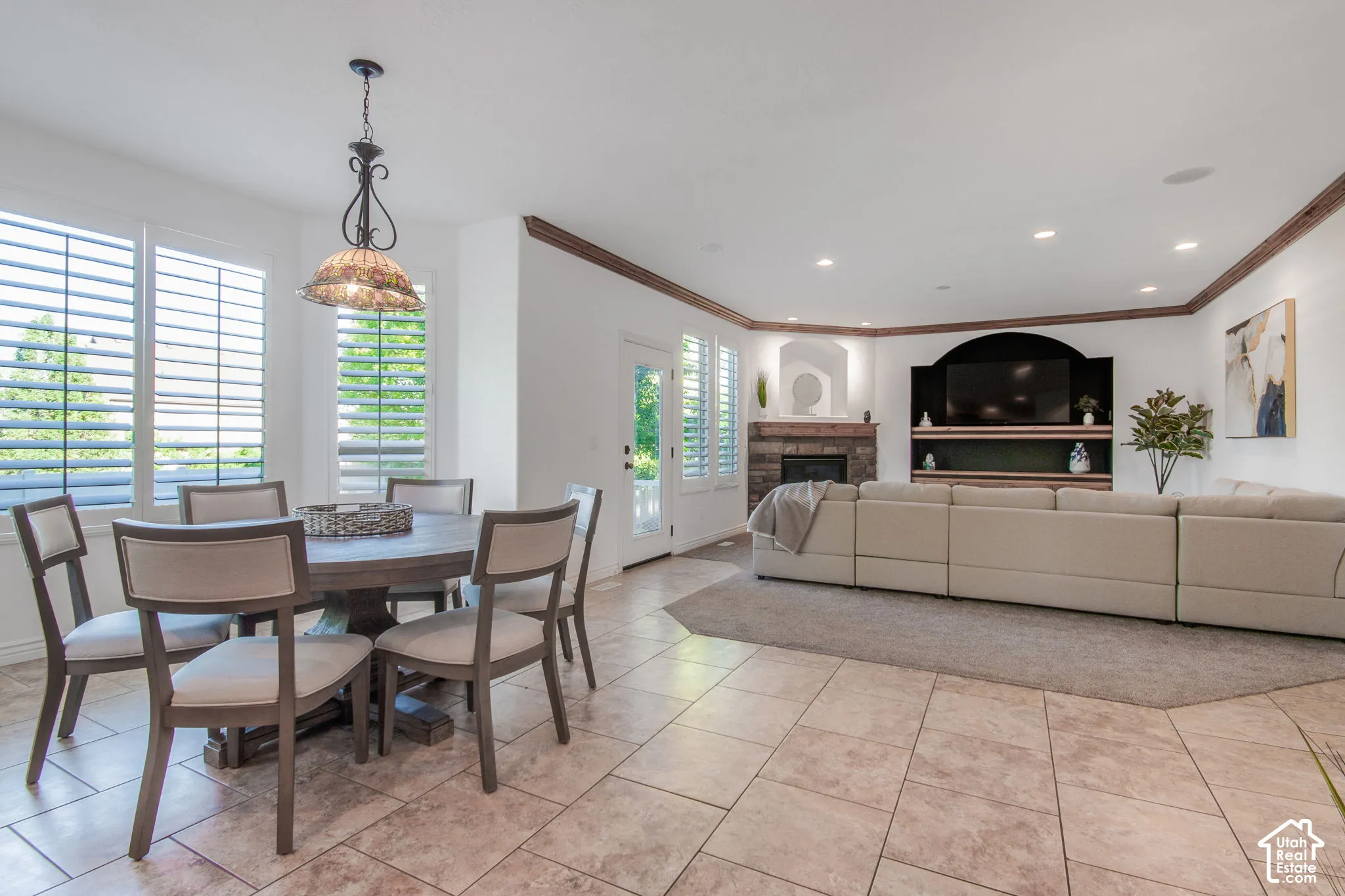Open floor plan featuring tile flooring, stone-surround fireplace, built-in entertainment cabinetry with arched detail, decorative crown molding, and plantation shutters