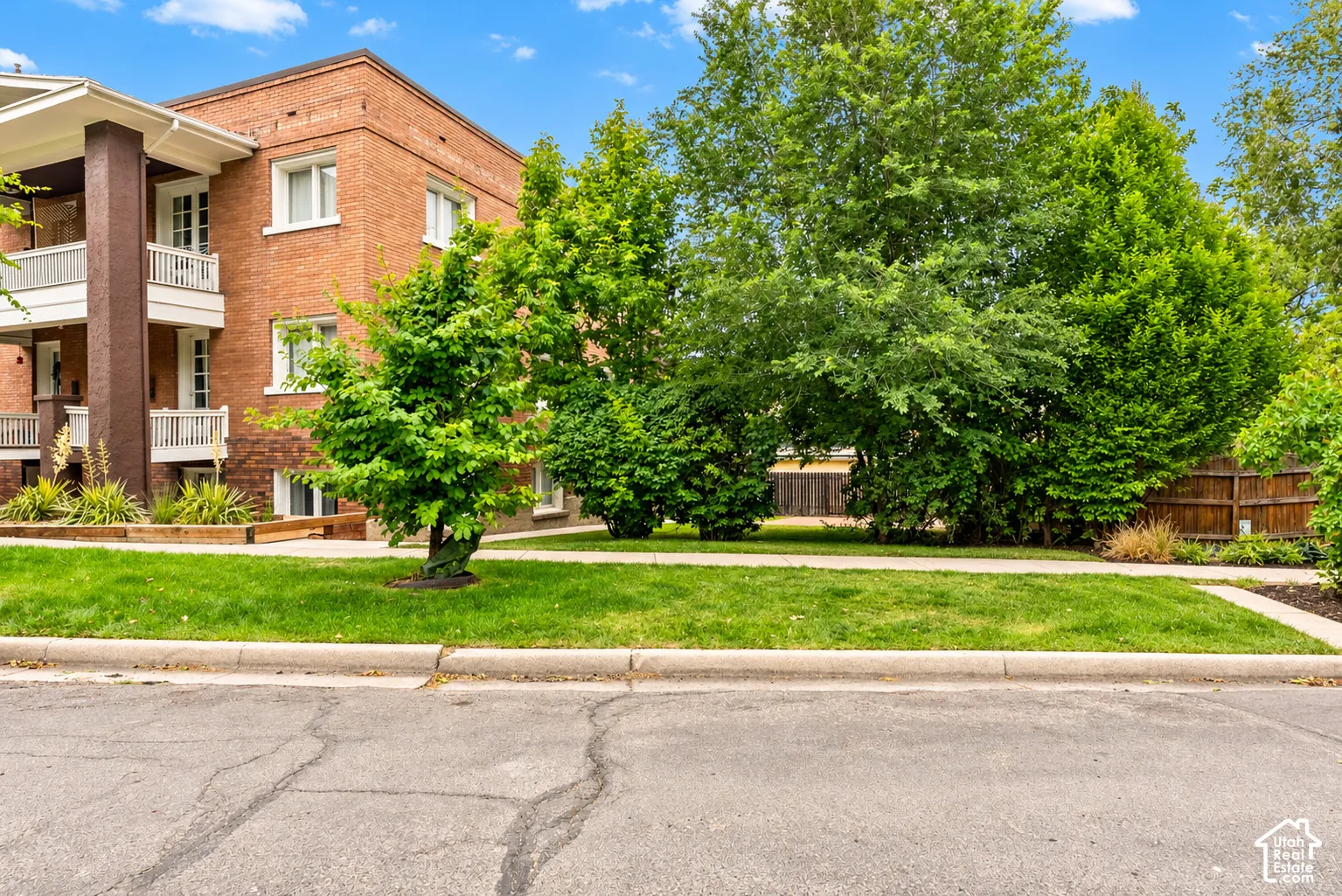 Multi-story brick exterior featuring tiered balconies with white railings, concrete sidewalk, and mature landscaping