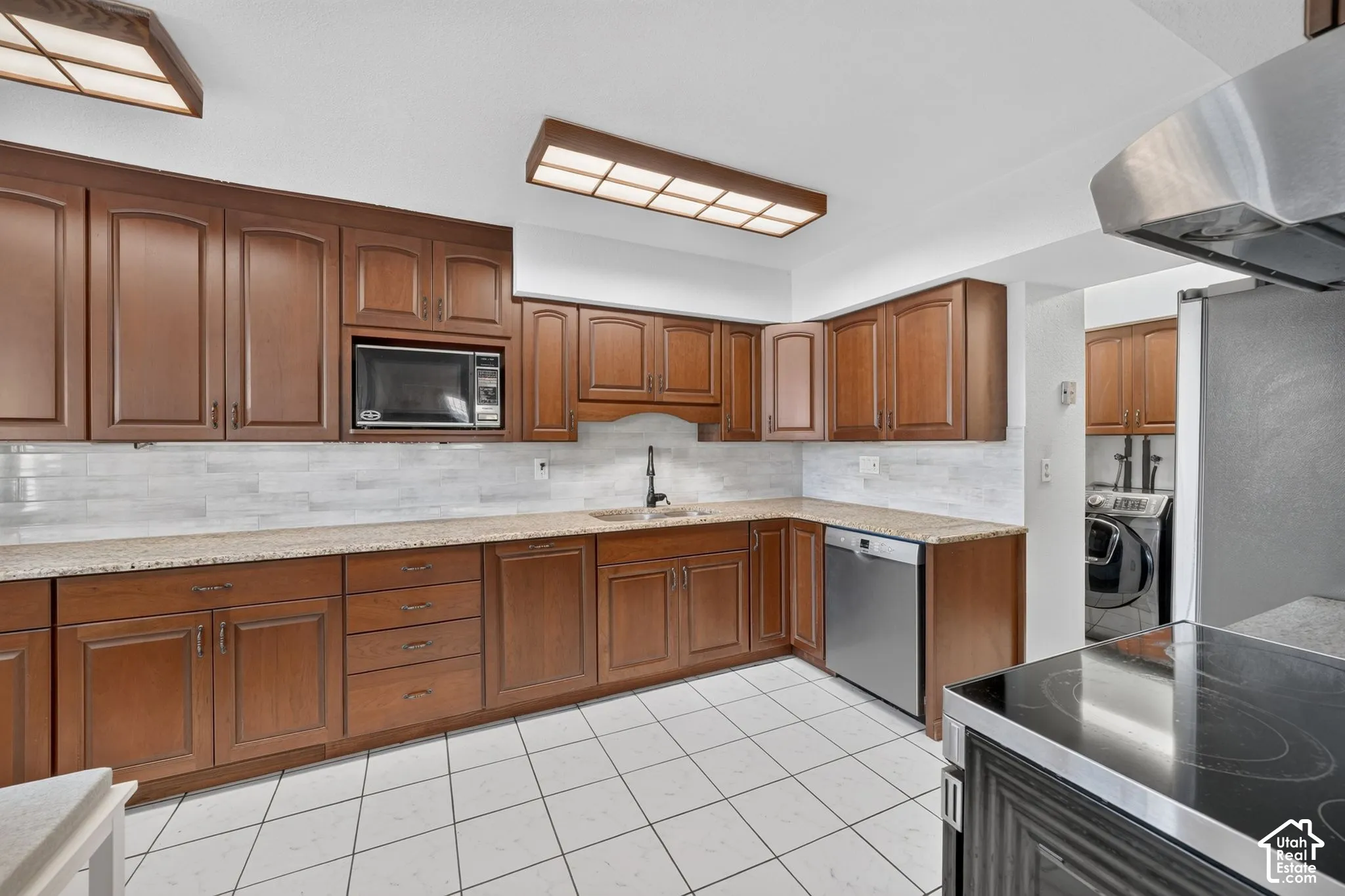 Kitchen featuring stained wood cabinetry, stone-finish countertops, tiled backsplash, stainless steel dishwasher, and ceramic tile flooring