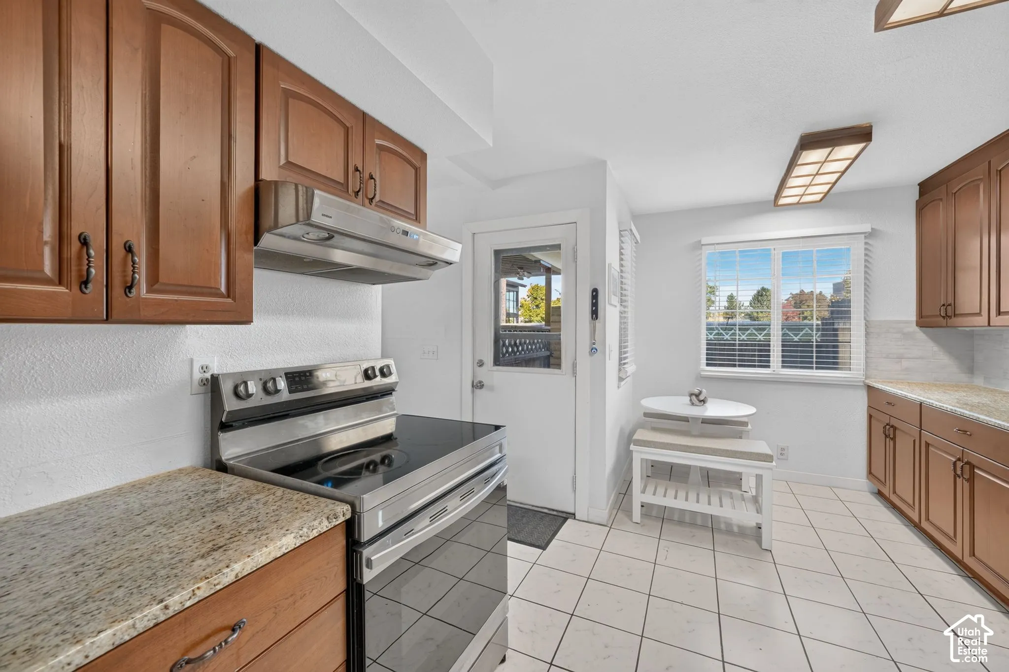 Kitchen featuring wood cabinetry, stone countertops, stainless steel range with overhead vent hood, ceramic tile flooring, and a glass-paneled exterior door