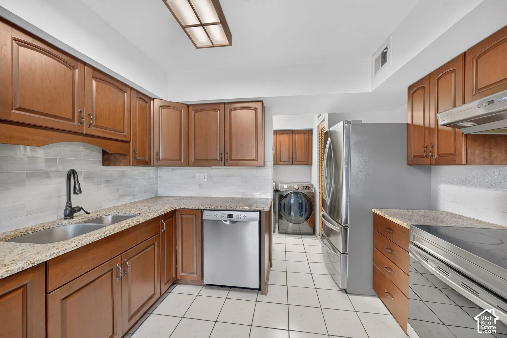 Kitchen featuring stained wood cabinetry, stone countertops, tiled backsplash, stainless steel appliances, and tile flooring