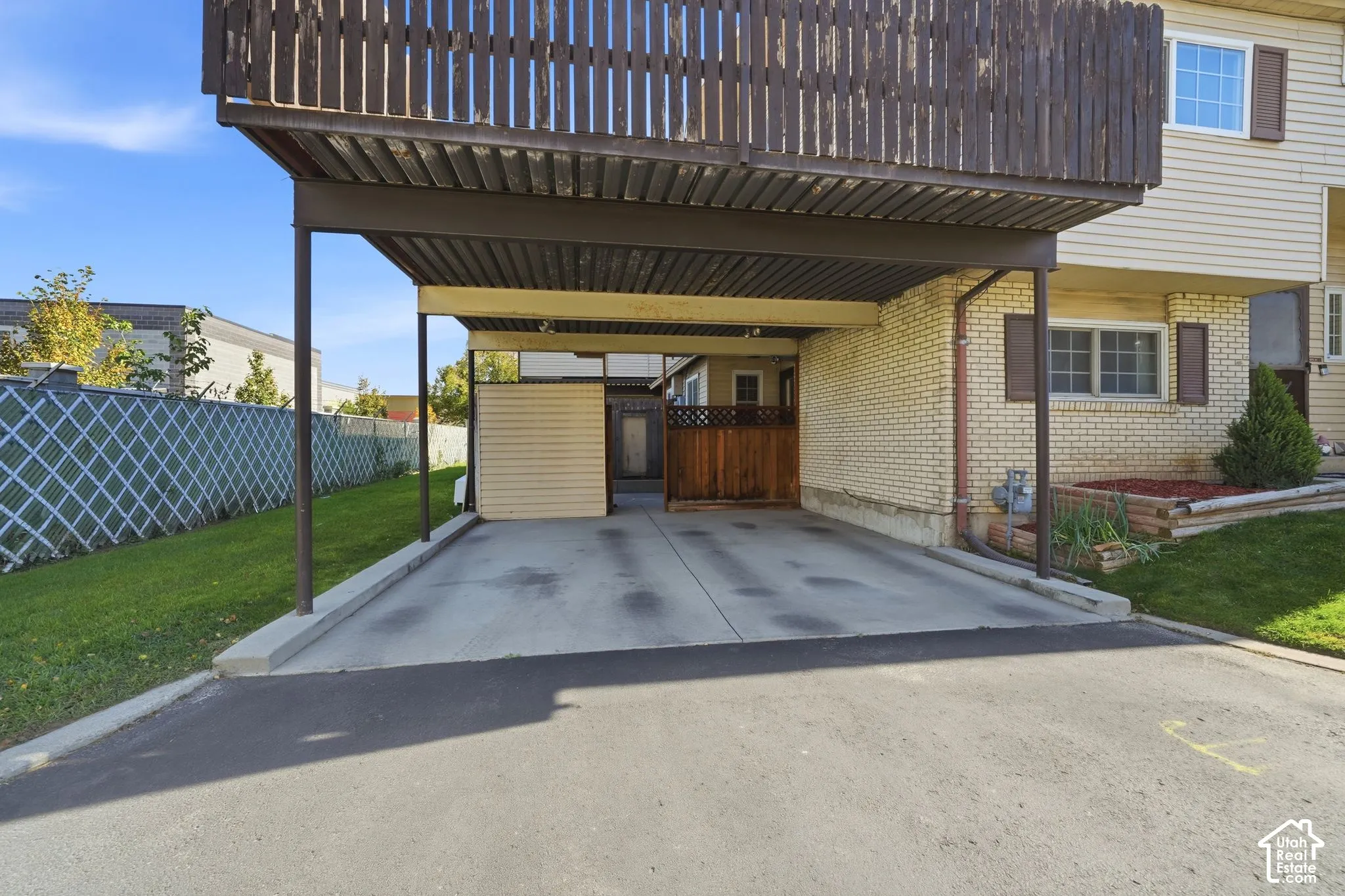 Covered carport with concrete flooring, attached balcony featuring vertical wood-slat privacy screening, light-colored brick masonry, and perimeter lattice fencing