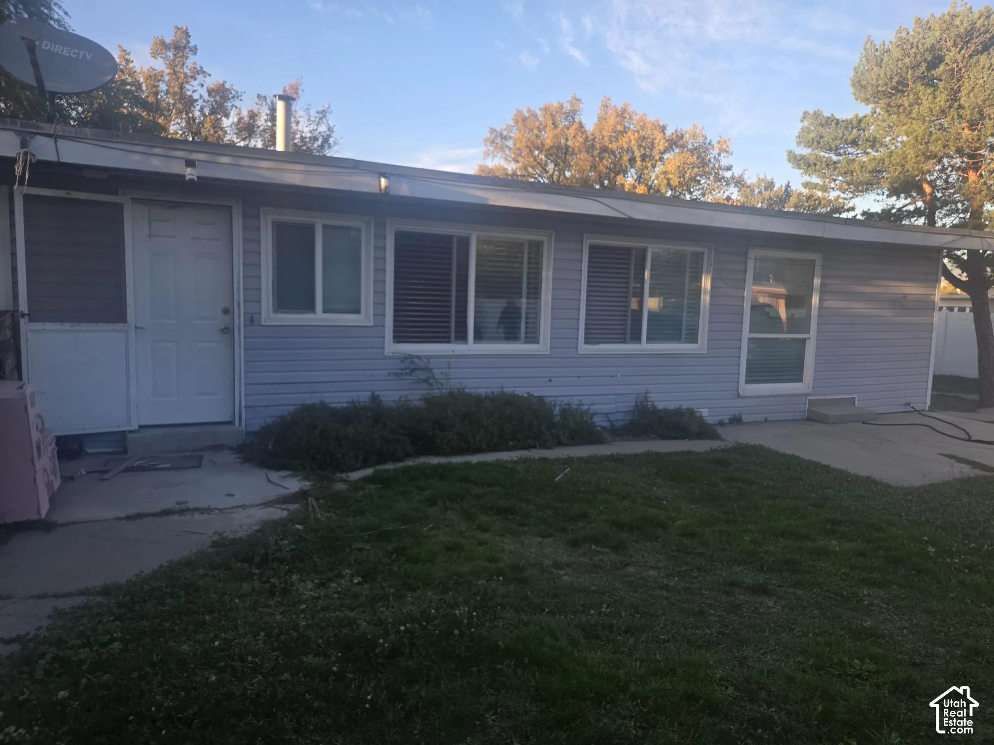 Single-story residence featuring light-colored horizontal siding, multiple sliding windows, a concrete patio, and mature landscaping