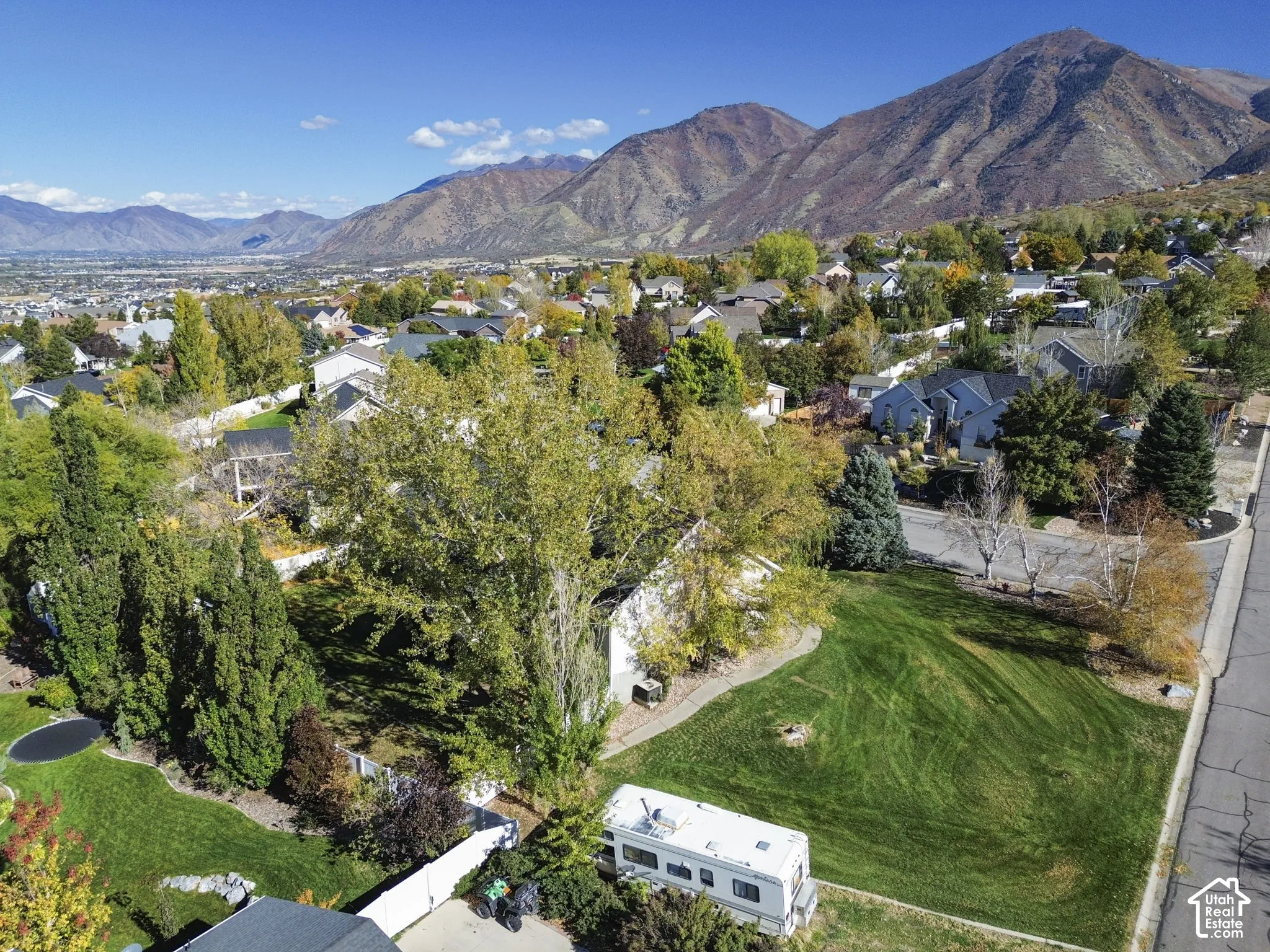 Aerial view of a residential lot featuring a expansive grass lawn, mature deciduous trees, evergreen foliage, and a prominent mountain backdrop