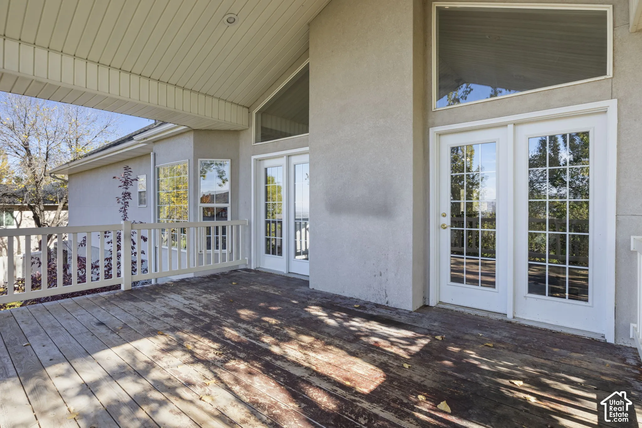 Covered exterior deck featuring wood decking, white balustrade railing, dual sets of multi-light glass doors, and expansive trapezoidal clerestory windows