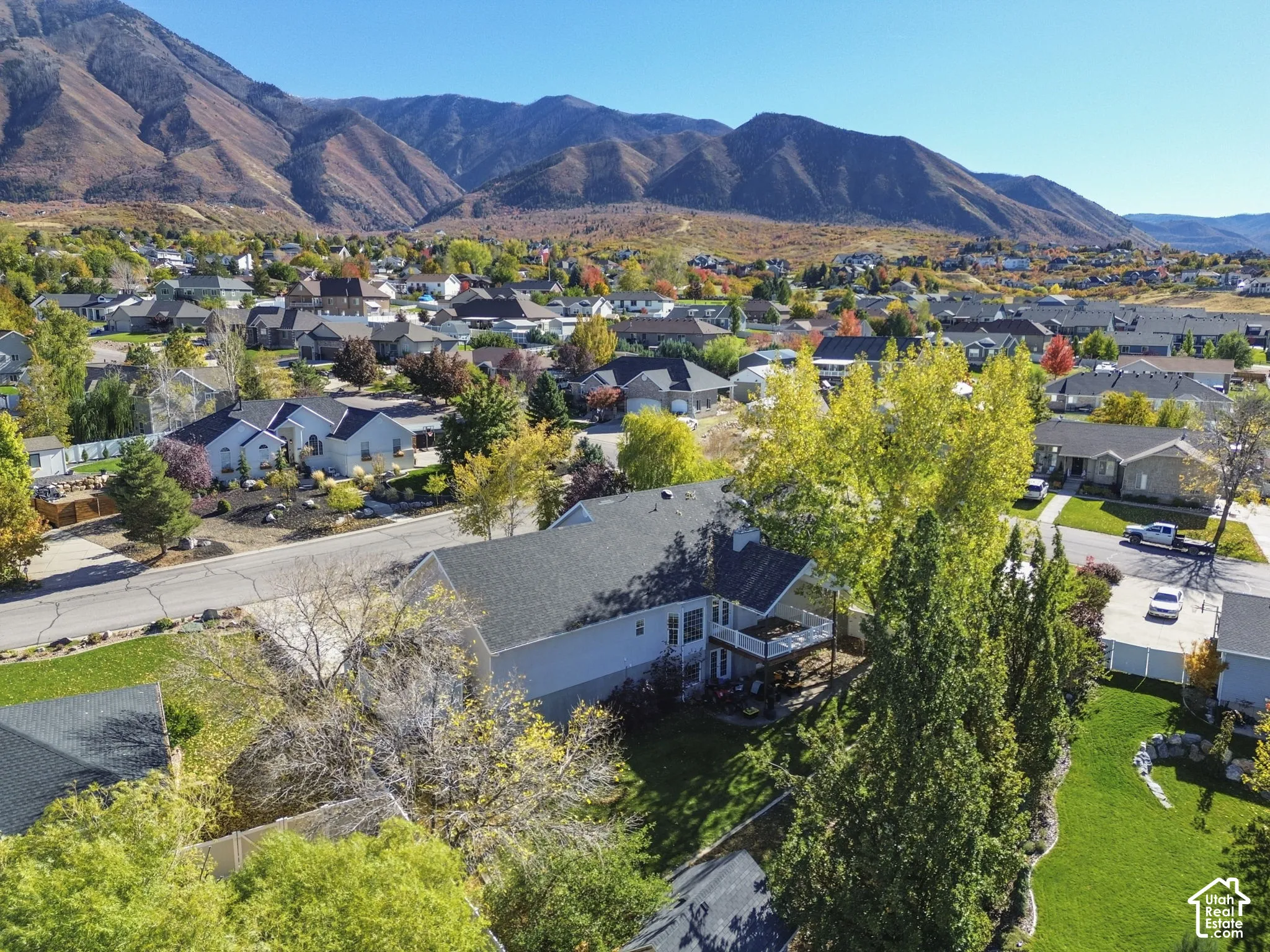 Aerial perspective featuring a multi-level residence with a raised deck, expansive lawn, mature perimeter trees, and a mountainous backdrop