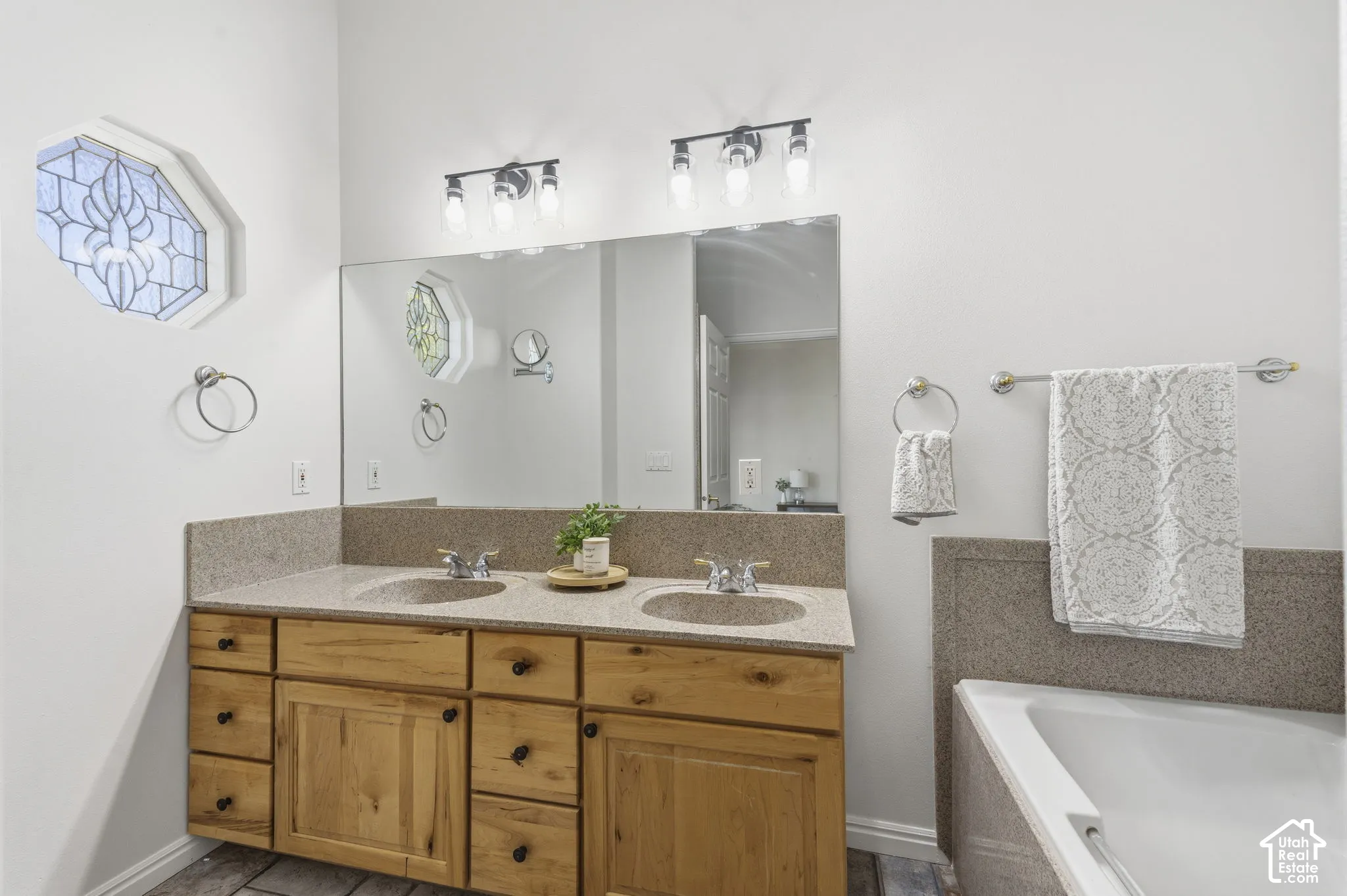 Dual-sink vanity with wood cabinetry, solid-surface countertops, expansive wall-mounted mirror, decorative geometric accent window, and wall-mounted light fixtures