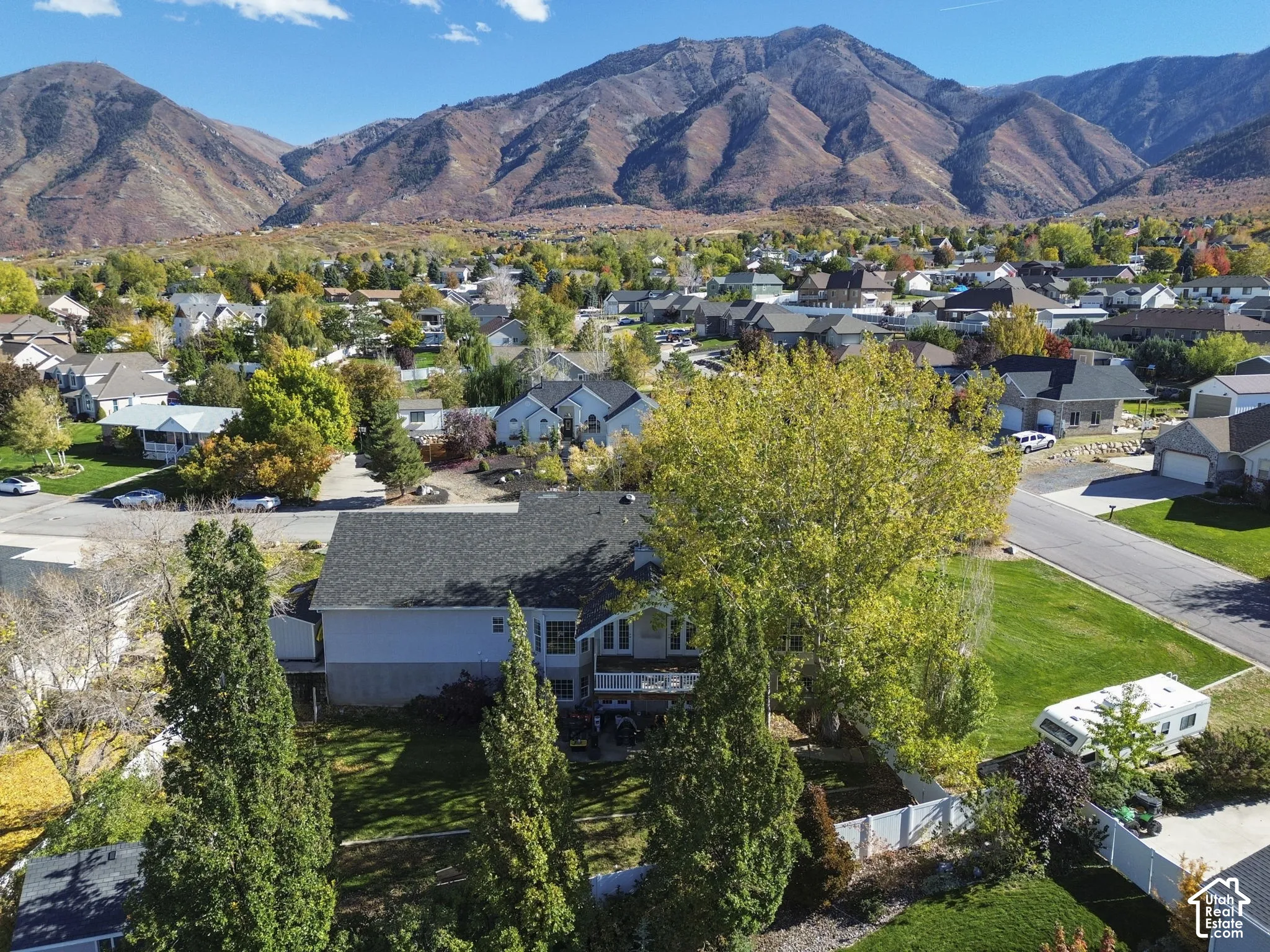 Aerial perspective of a multi-level residence featuring a dark shingle roof, rear deck, expansive lawn, mature landscaping, and prominent mountain backdrop