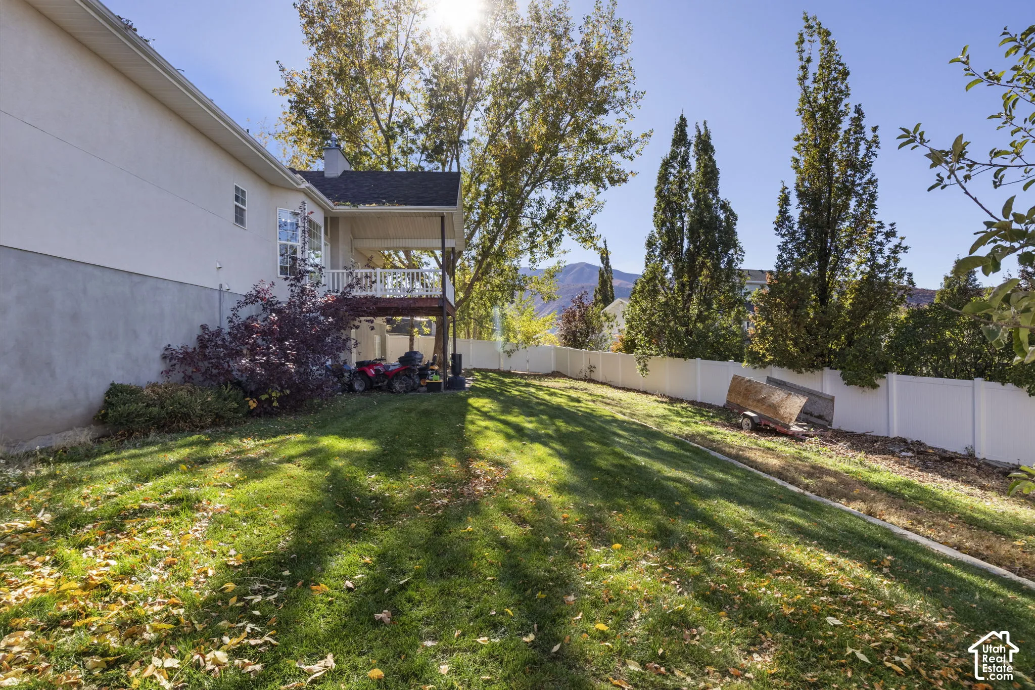 Backyard featuring a covered deck, white vinyl perimeter fencing, mature trees, and a mountain backdrop