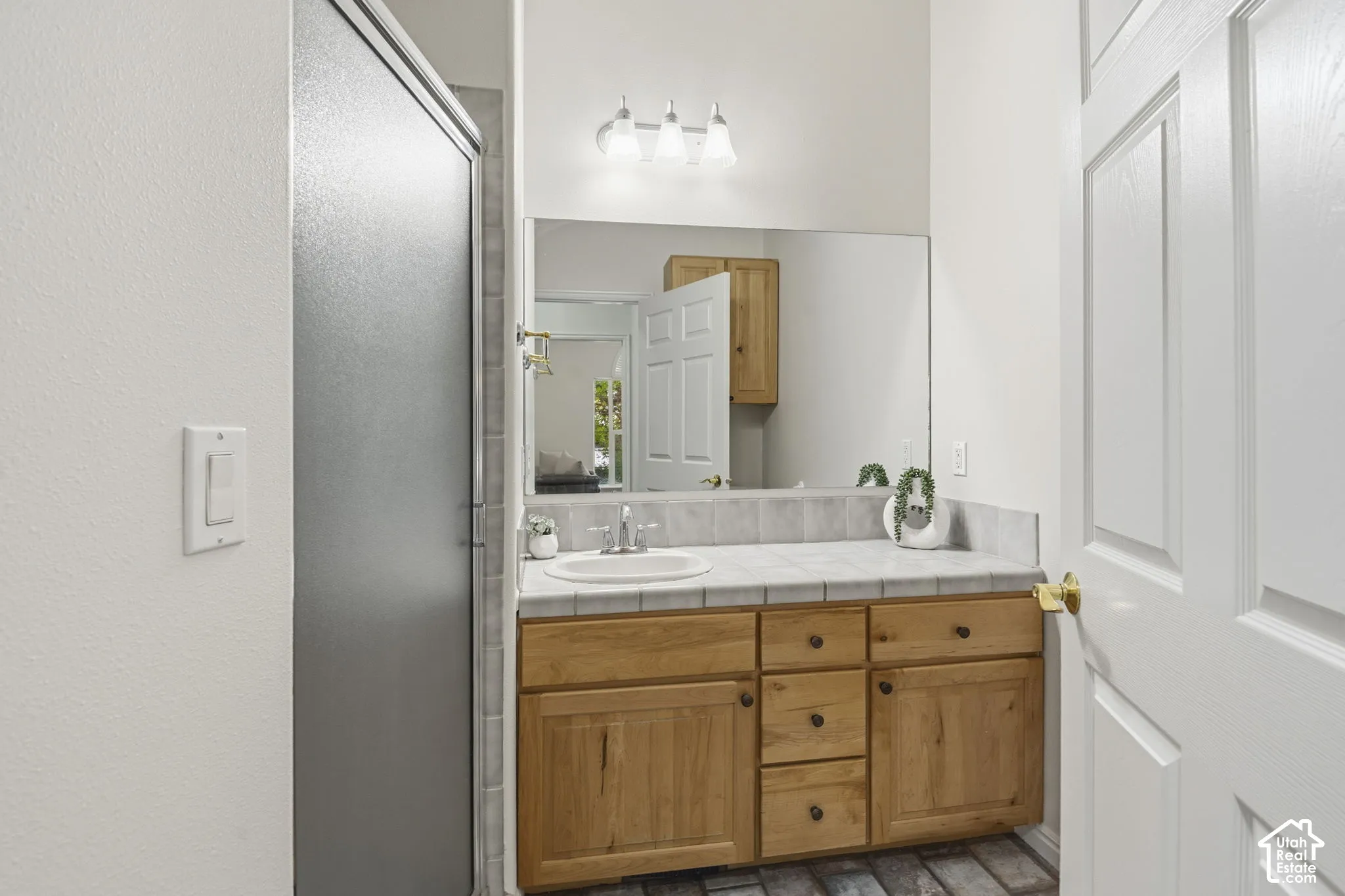 Bathroom featuring a wood-finish vanity with a tile countertop, integrated basin, oversized wall-mounted mirror, and three-bulb vanity lighting