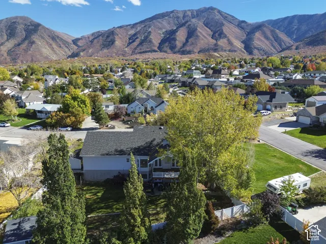 Aerial perspective of a multi-level residence featuring a dark shingle roof, rear deck, expansive lawn, mature landscaping, and prominent mountain backdrop