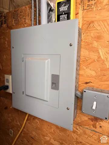 Electrical service panel featuring a metal enclosure mounted on oriented strand board (OSB) wall surfacing, inclusive of an exterior-mounted light switch and a standard grounded power outlet