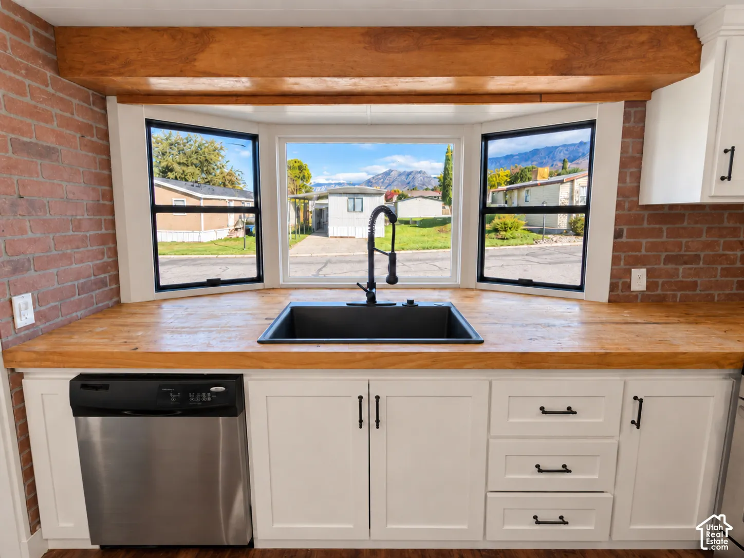 Kitchen area featuring a wood-finish countertop, exposed brick walls, matte black faucet with an integrated undermount sink, custom white cabinetry, and a wood-beam ceiling accent