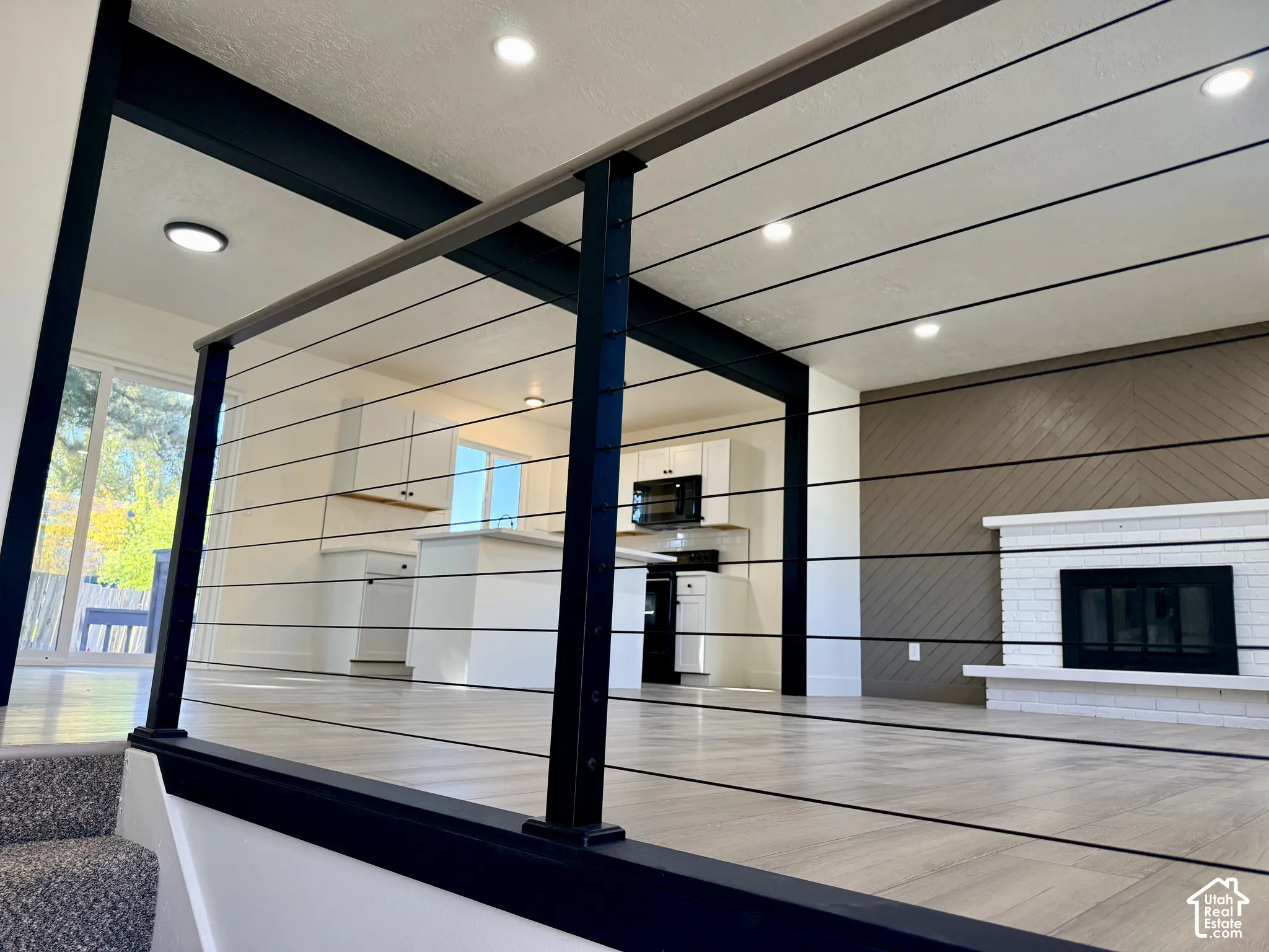Modern horizontal cable railing with matte black posts, white brick fireplace, diagonal wood-patterned accent wall, recessed lighting, and wood-finish flooring