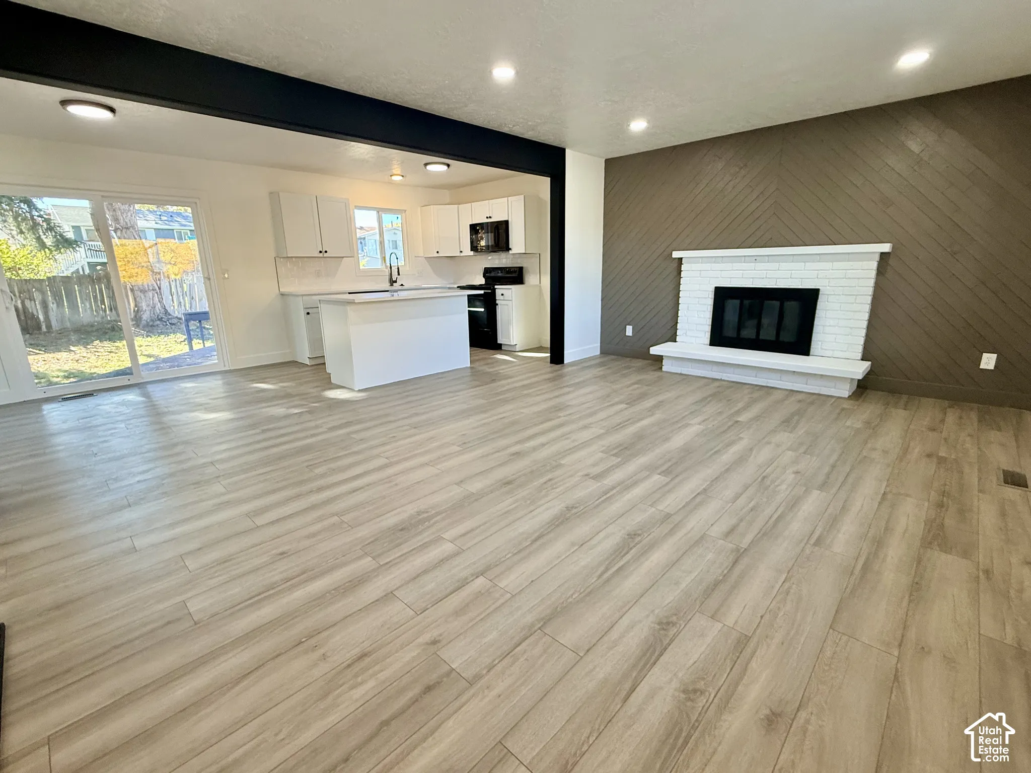 Wood-finish flooring, white brick fireplace, herringbone-patterned accent wall, kitchen island, and recessed lighting